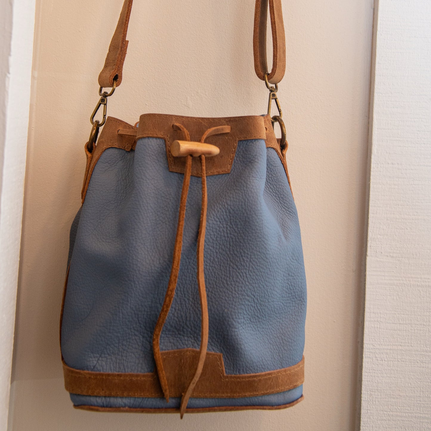 A blue pebbled leather bucket bag with tan leather trim, drawstring closure, and shoulder strap, hanging against a light beige wall.
