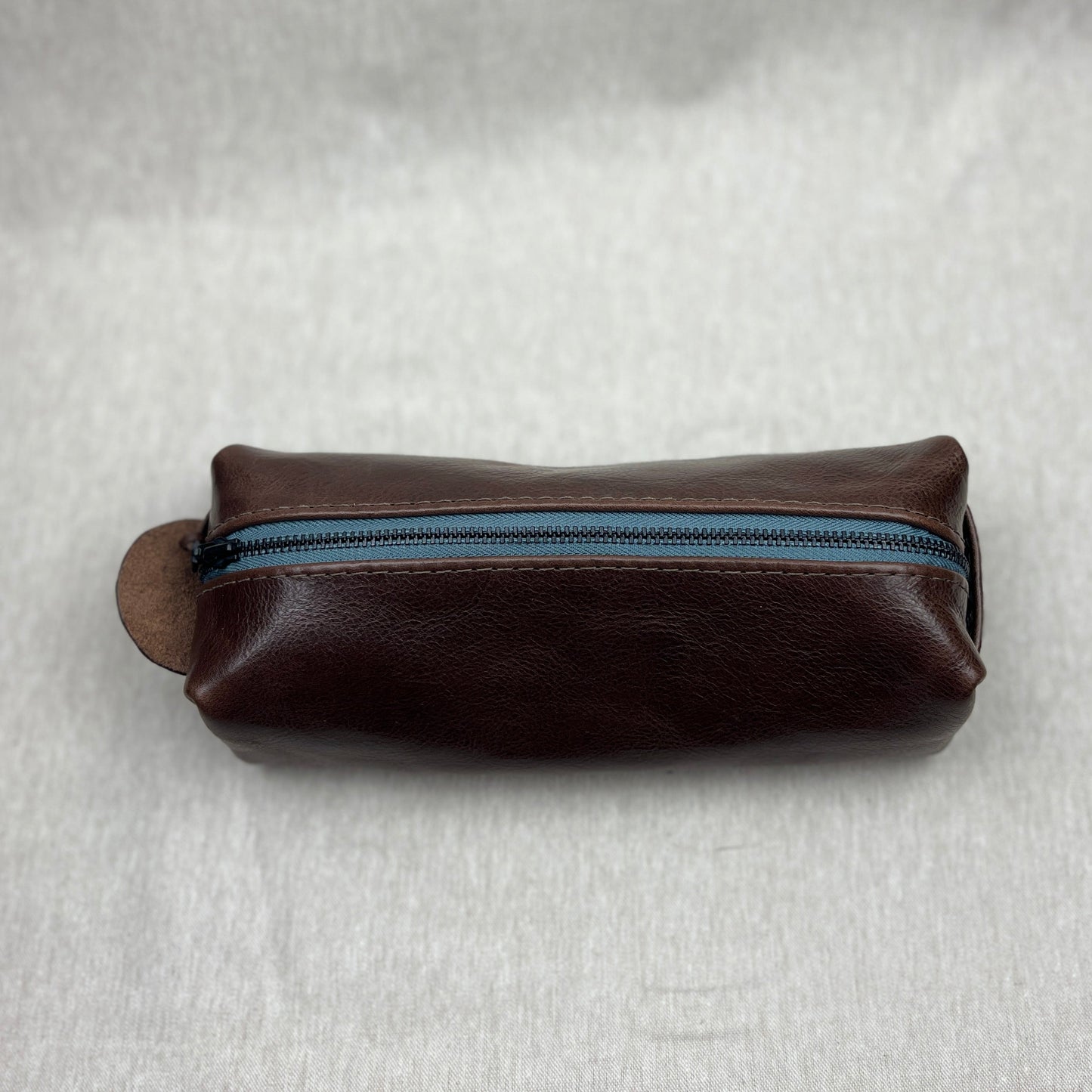 A dark brown leather zippered pouch with a blue zipper track, resting on a textured light gray fabric background.