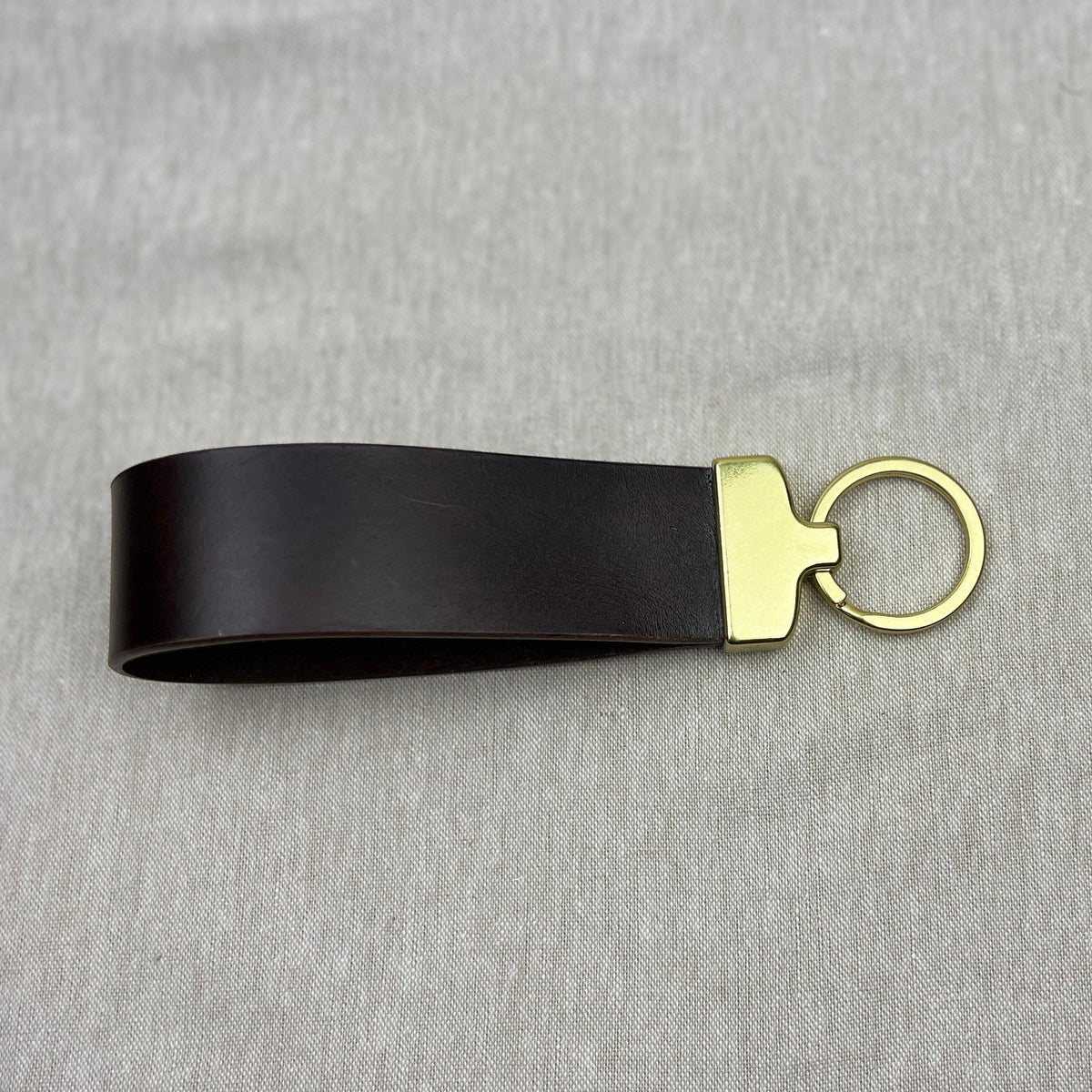 A black leather keychain strap with a gold-tone metal connector and split key ring, displayed on a textured gray fabric background.
