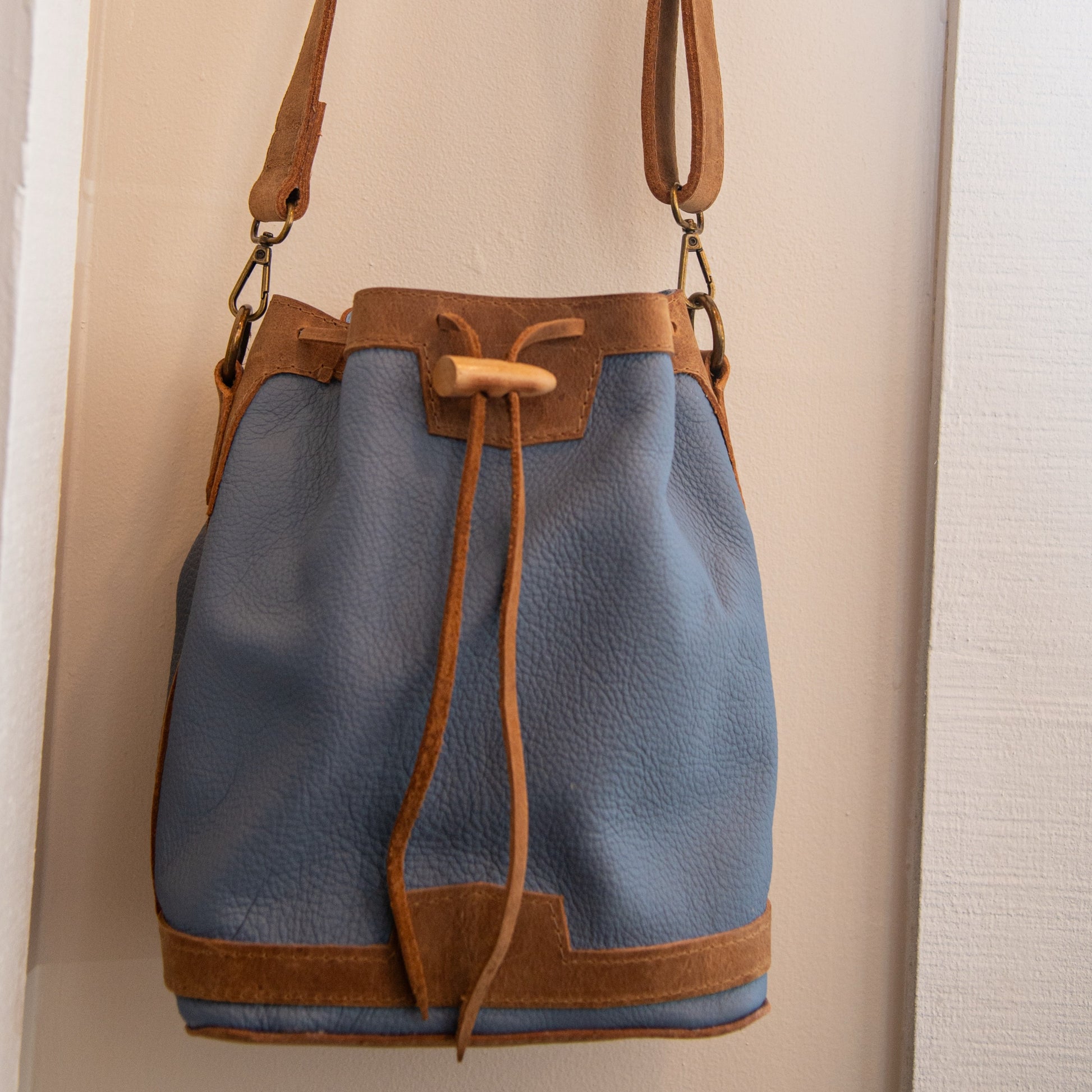 A blue pebbled leather bucket bag with tan leather trim, drawstring closure, and shoulder strap, hanging against a light beige wall.