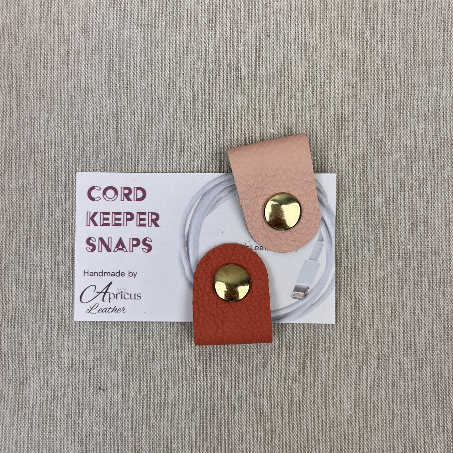 Two cord keepers with gold-tone snaps, one in apricot and one in blush pink, displayed on a white backing card over a textured beige fabric background. 