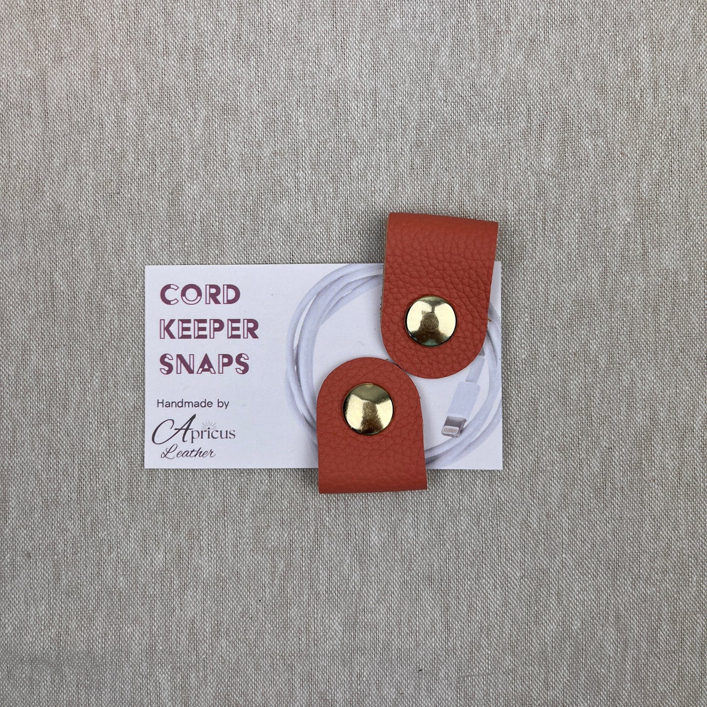 A pair of salmon leather cord keeper snaps with gold-tone metal studs, presented on a white backing card over a textured gray fabric background.