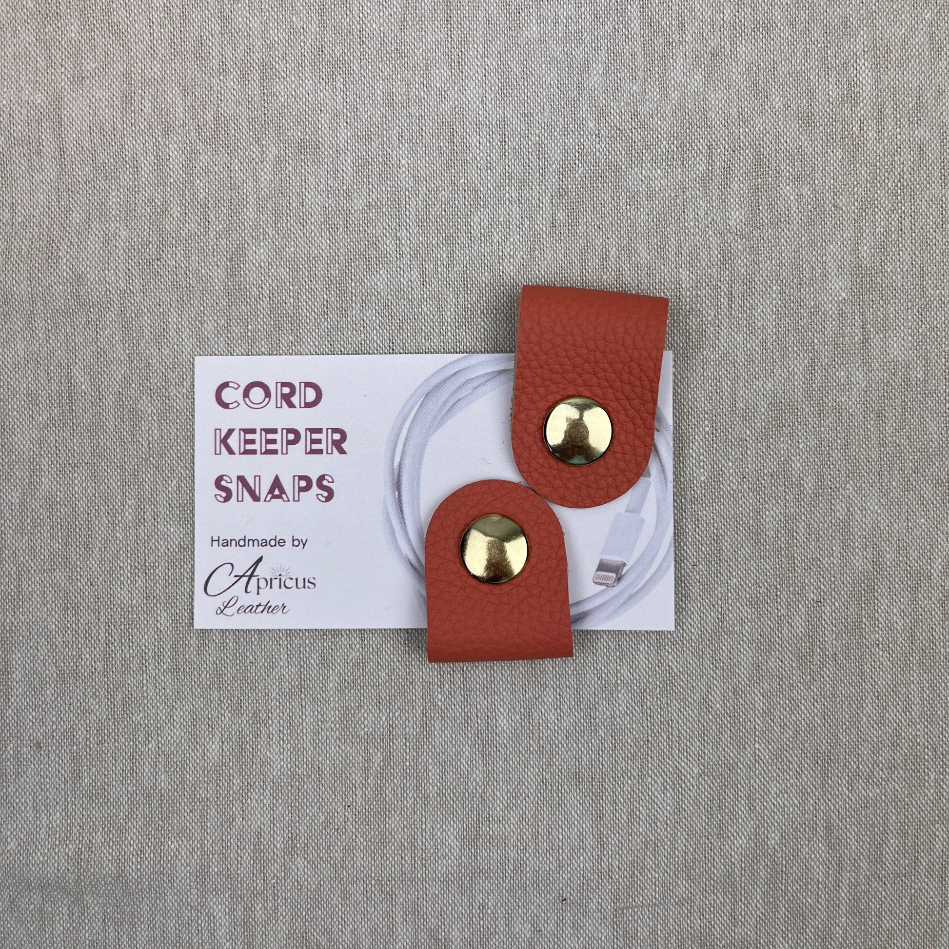 A pair of salmon leather cord keeper snaps with gold-tone metal studs, presented on a white backing card over a textured gray fabric background.