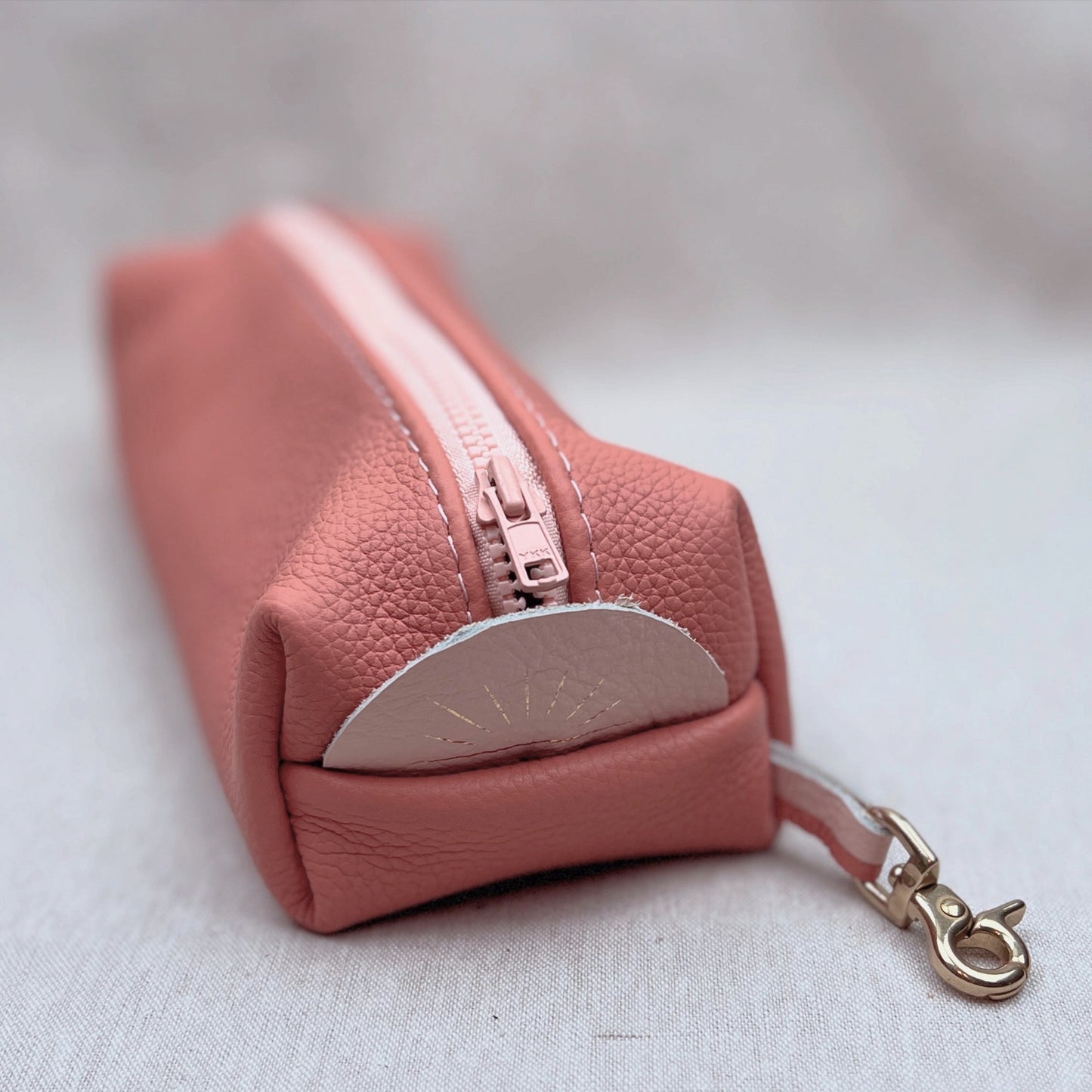 A dusty pink textured leather zip pouch with a silver metal clasp and a light pink zipper pull, resting on a light gray fabric surface.