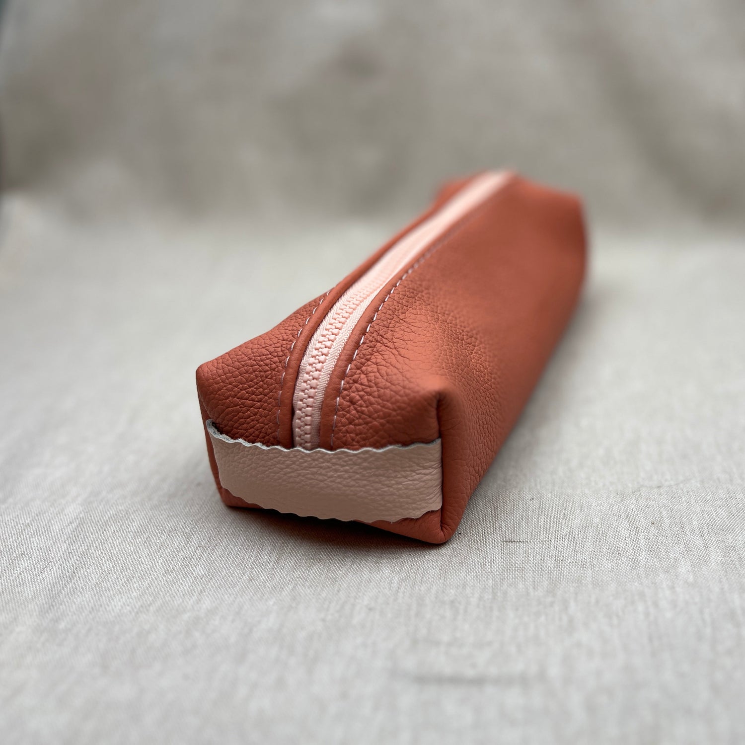 An apricot pink leather zippered pouch with a light pink zipper and a matching pink fabric tab at the base, resting on a textured light gray surface.