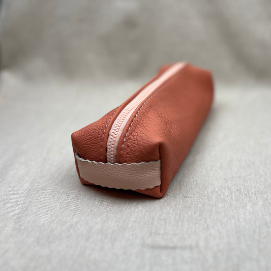 An apricot pink leather zippered pouch with a light pink zipper and a matching pink fabric tab at the base, resting on a textured light gray surface.