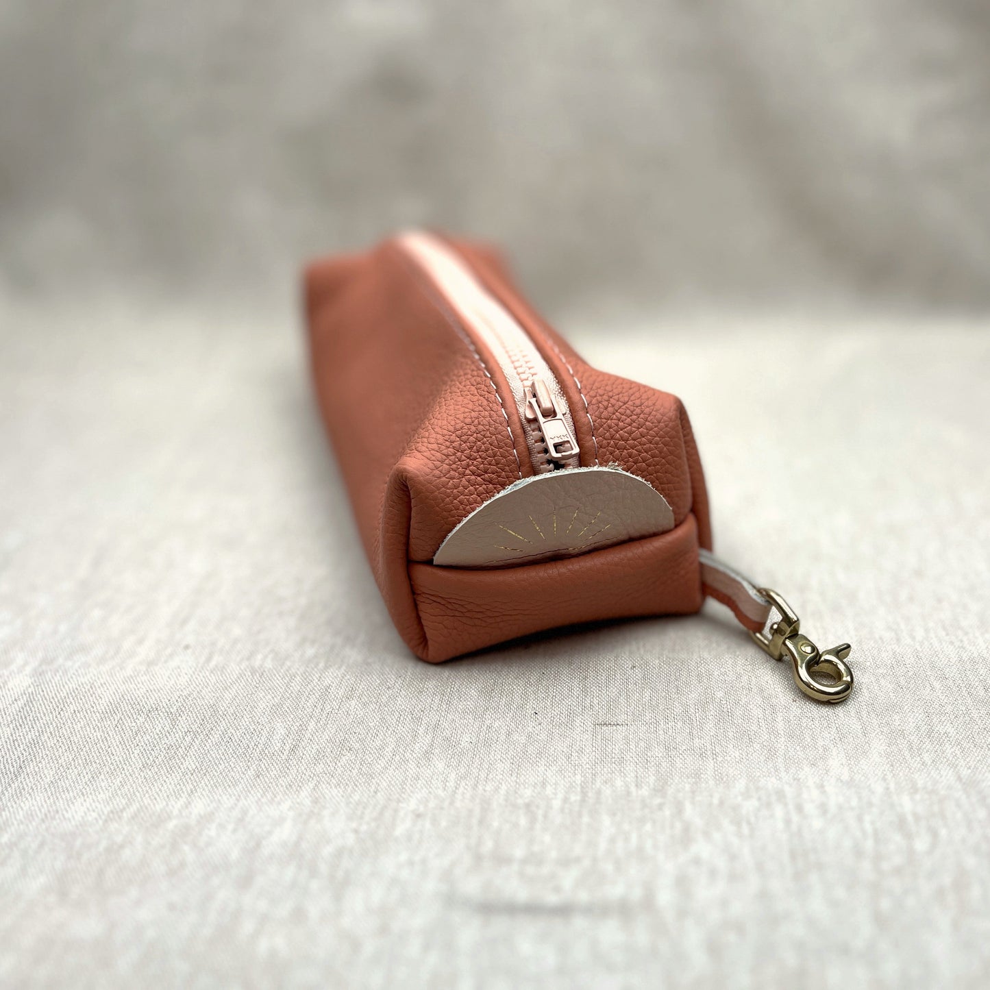 A small pebbled leather pouch in a warm terracotta pink color, featuring a light pink zipper, a leather accent tab, and a brass metal snap hook clasp, resting on a textured light gray fabric surface.