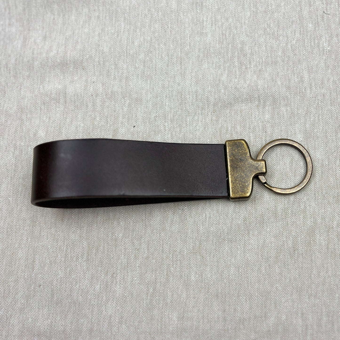 A black leather keychain strap with an antique gold-tone metal connector and split key ring, displayed on a textured gray fabric background.