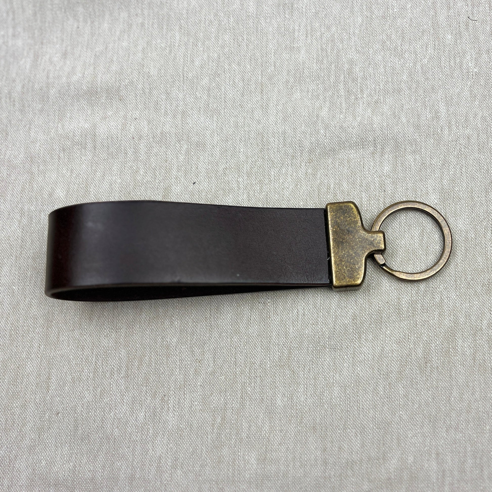 A black leather keychain strap with an antique gold-tone metal connector and split key ring, displayed on a textured gray fabric background.