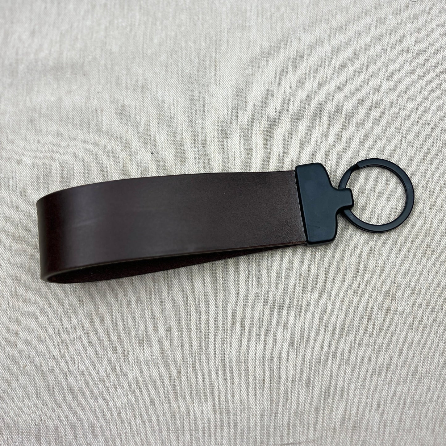 A dark brown leather strap keychain with a matte black metal connector and ring, displayed on a light gray textured fabric background.
