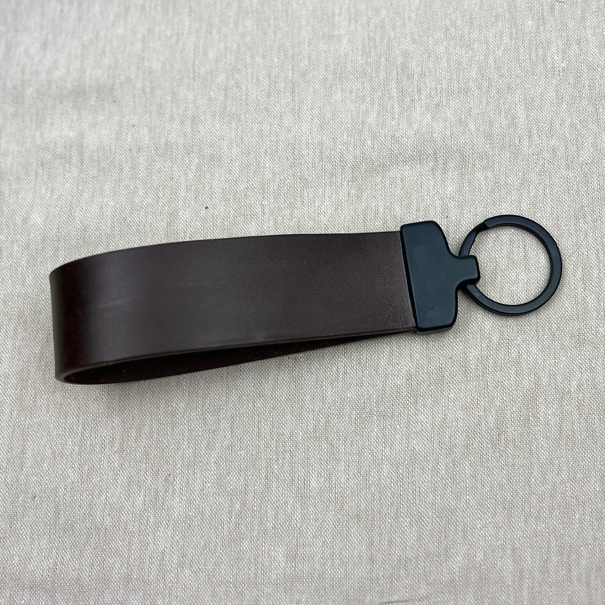 A dark brown leather strap keychain with a matte black metal connector and ring, displayed on a light gray textured fabric background.