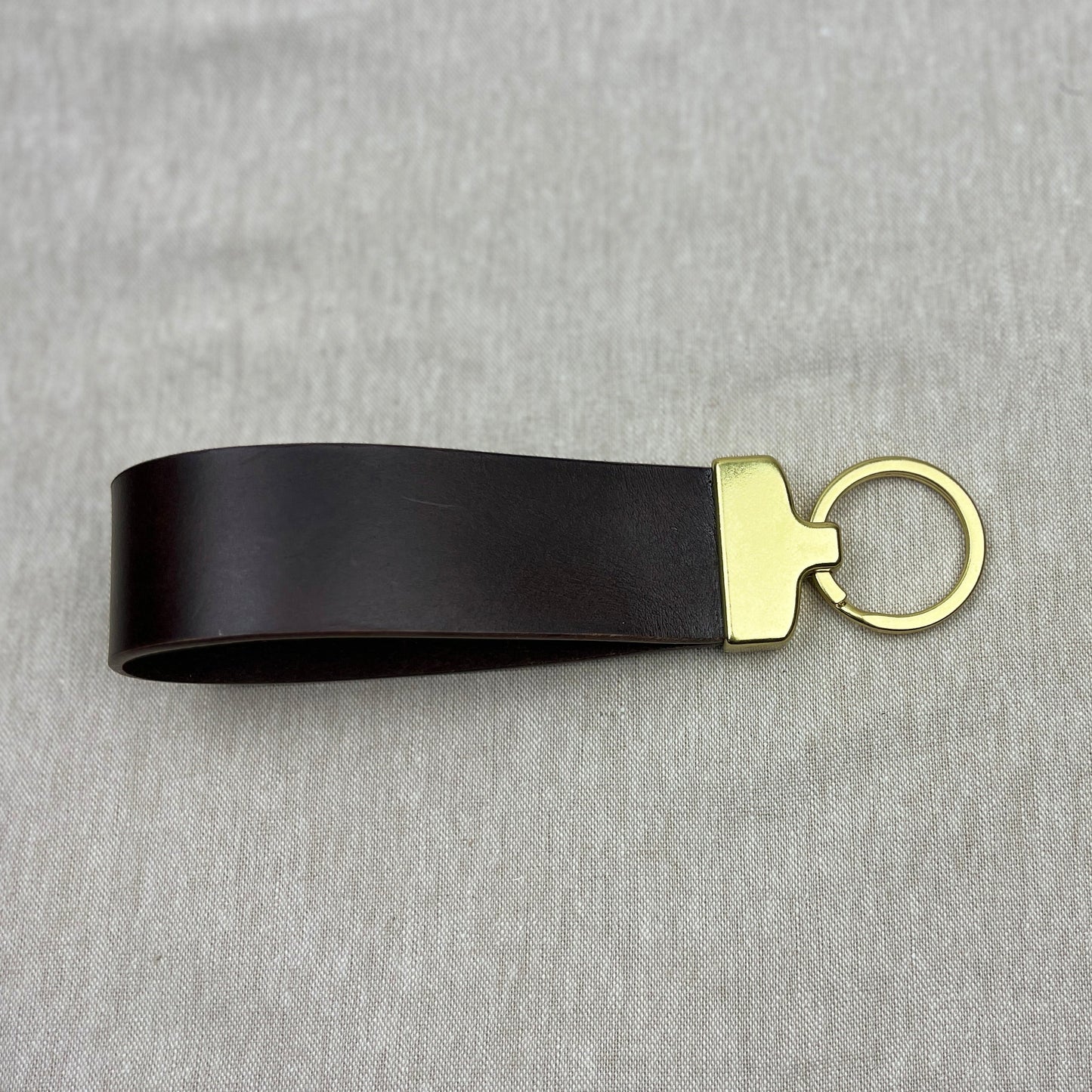 A black leather keychain strap with a gold-tone metal connector and split key ring, displayed on a textured gray fabric background.