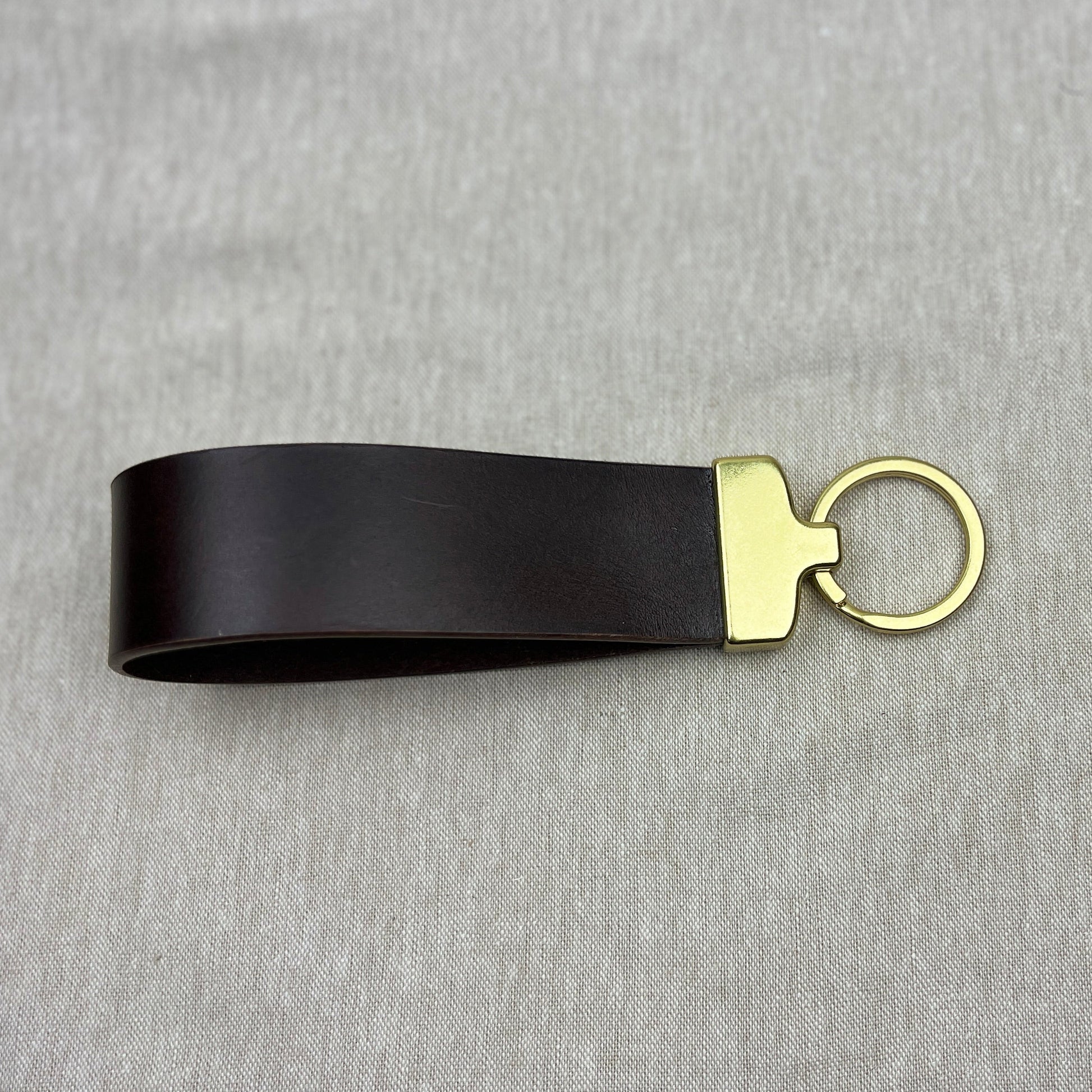 A black leather keychain strap with a gold-tone metal connector and split key ring, displayed on a textured gray fabric background.