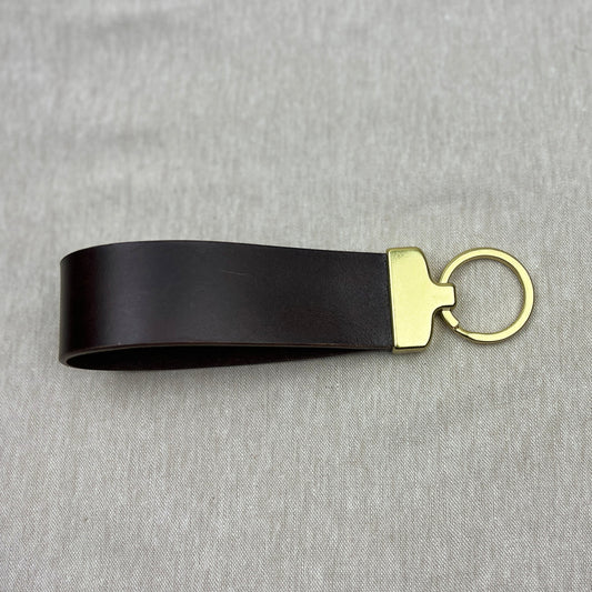A black leather keychain strap with a gold-tone metal connector and split key ring, displayed on a textured gray fabric background.