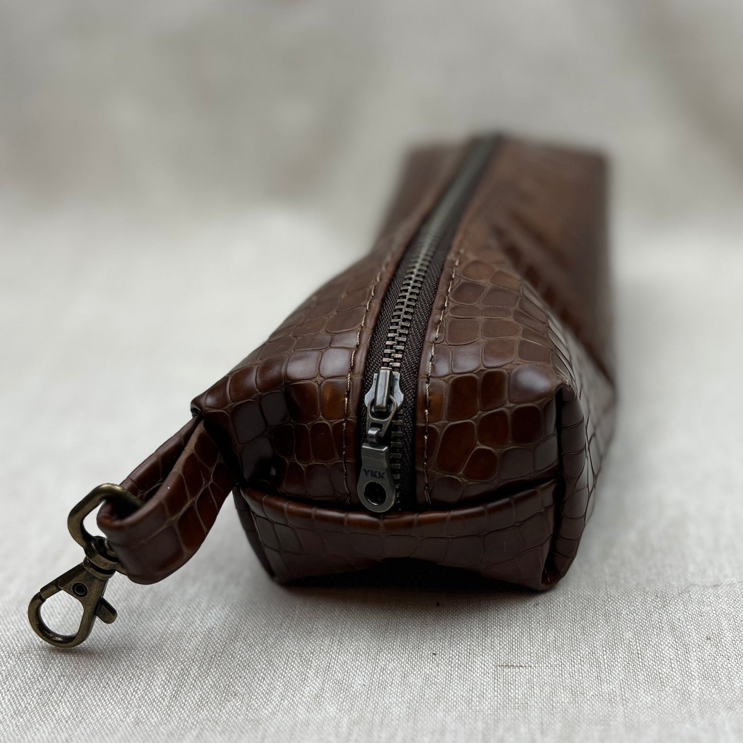 A brown crocodile-embossed leather zippered pouch with a matching wristlet strap and antique bronze clasp, resting on a textured light gray fabric surface.
