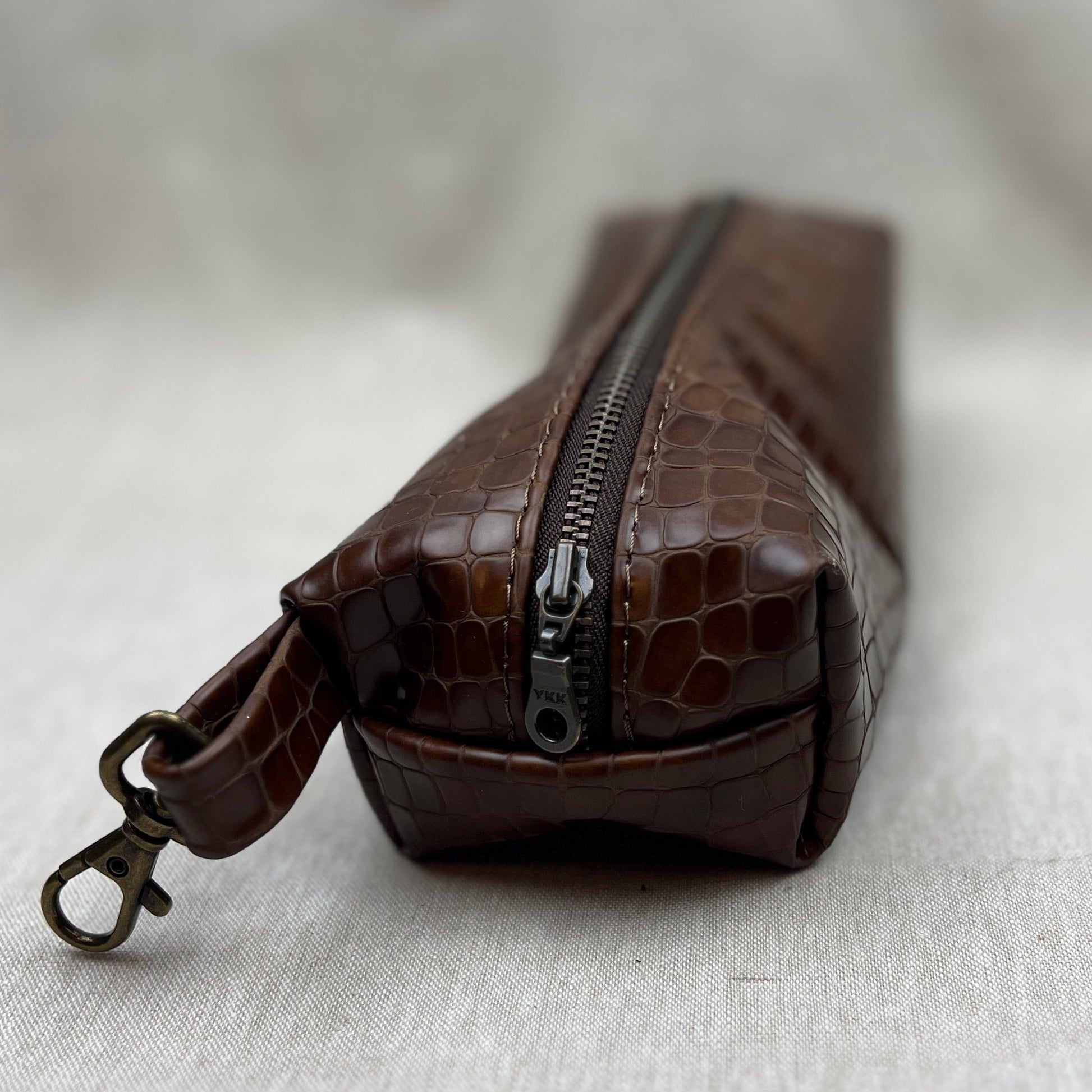 A brown crocodile-embossed leather zippered pouch with a matching wristlet strap and antique bronze clasp, resting on a textured light gray fabric surface.