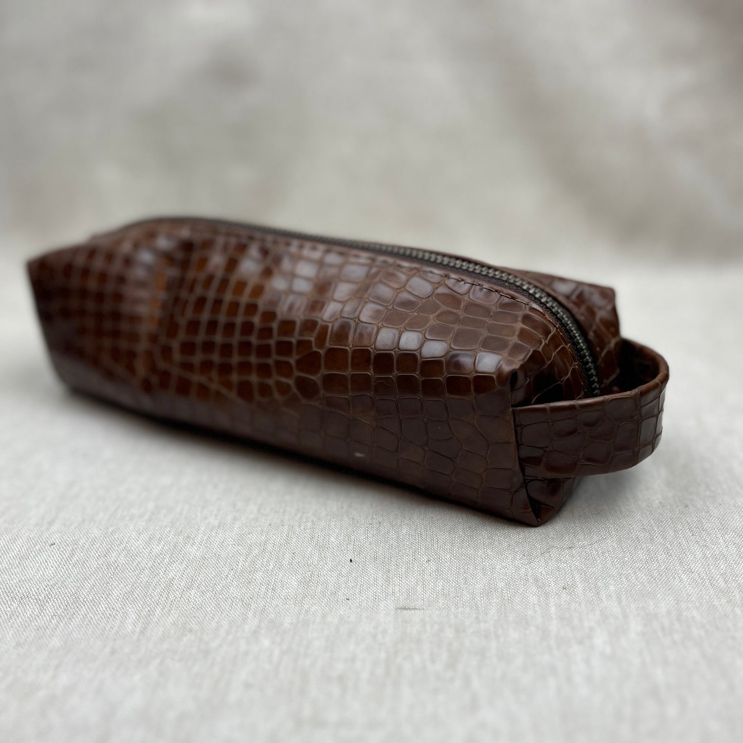 A brown crocodile-embossed leather zippered pouch with a matching leather loop handle, resting on a textured light gray fabric surface.