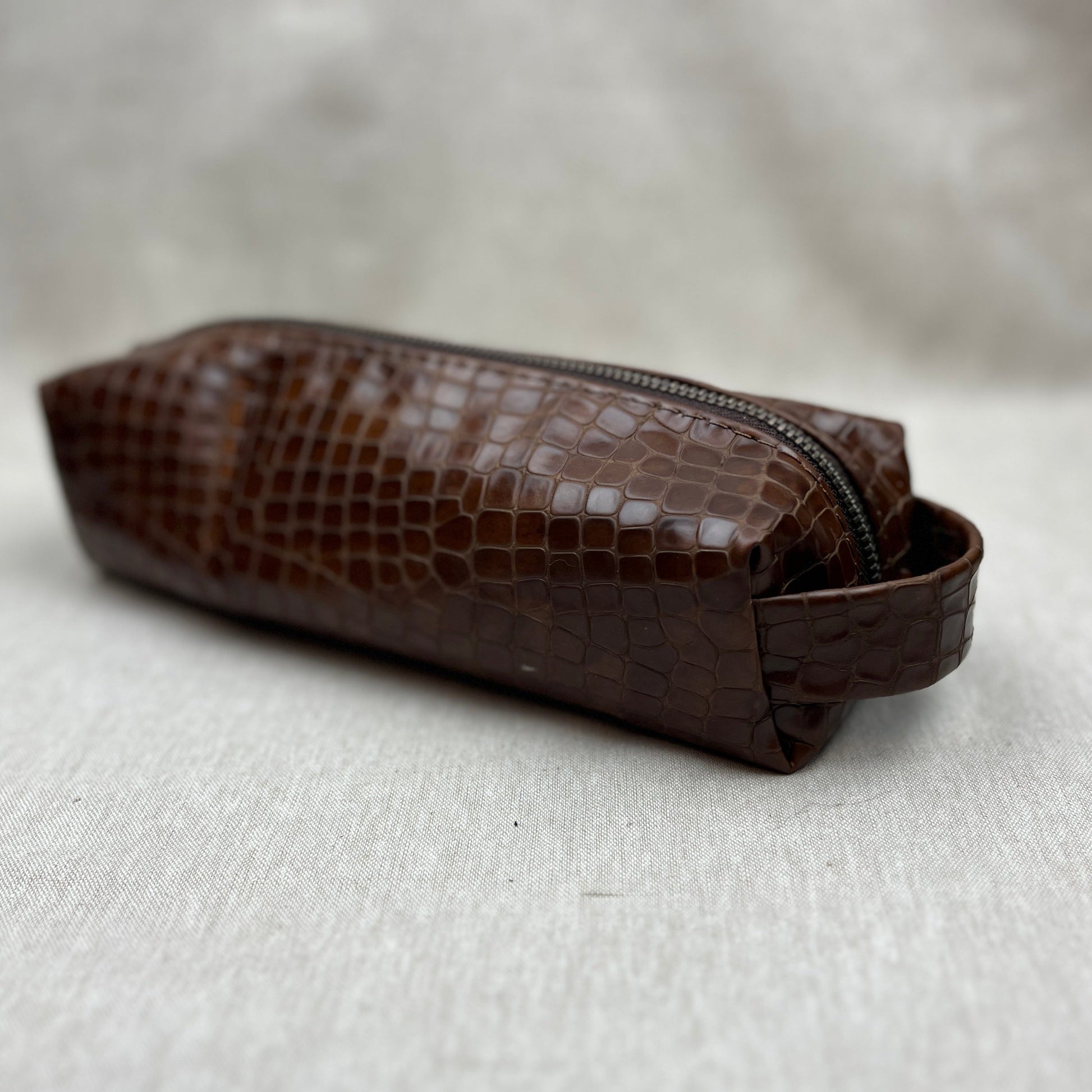 A brown crocodile-embossed leather zippered pouch with a matching leather loop handle, resting on a textured light gray fabric surface.