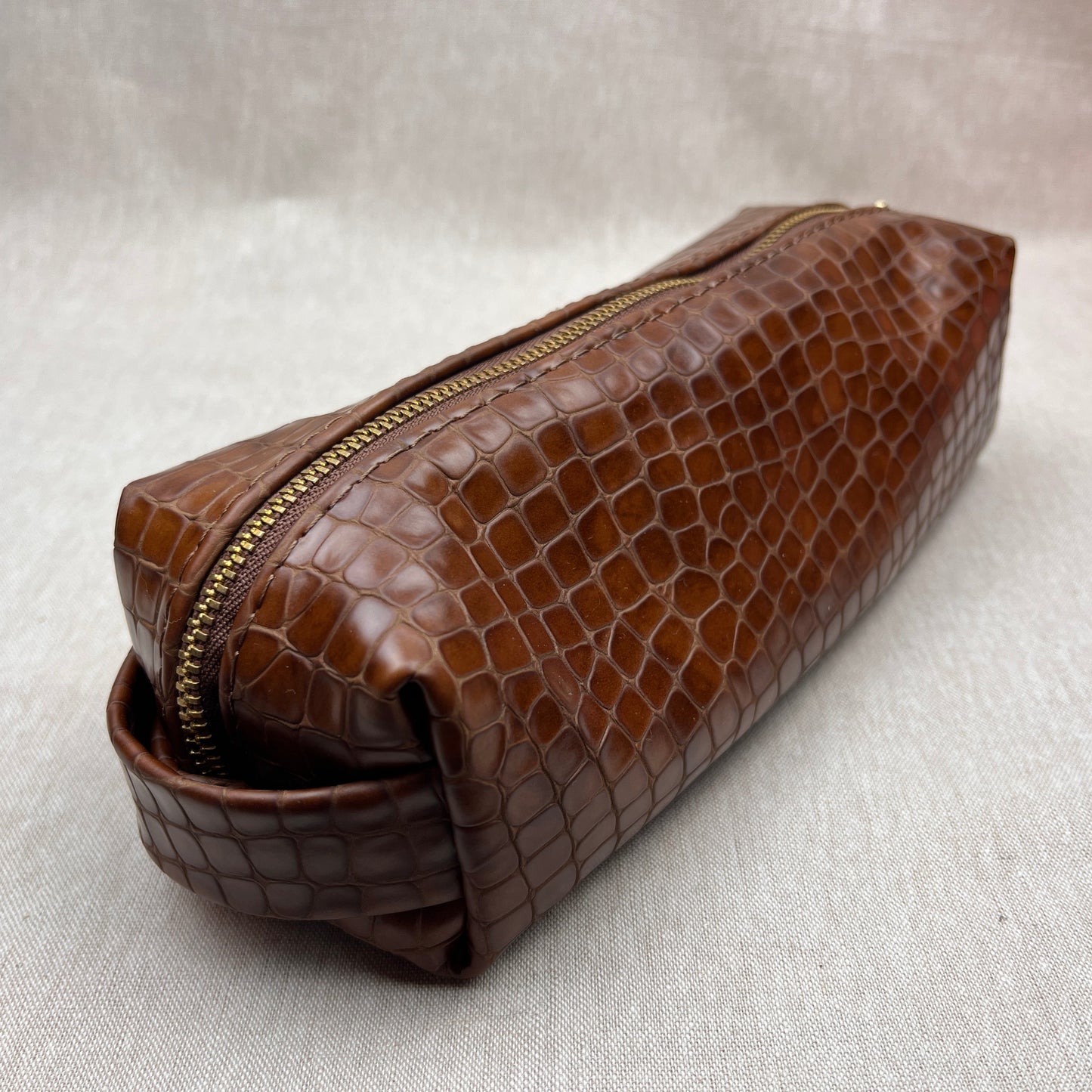 A brown crocodile-embossed leather zippered pouch with a matching leather loop handle, resting on a light beige textured fabric background.