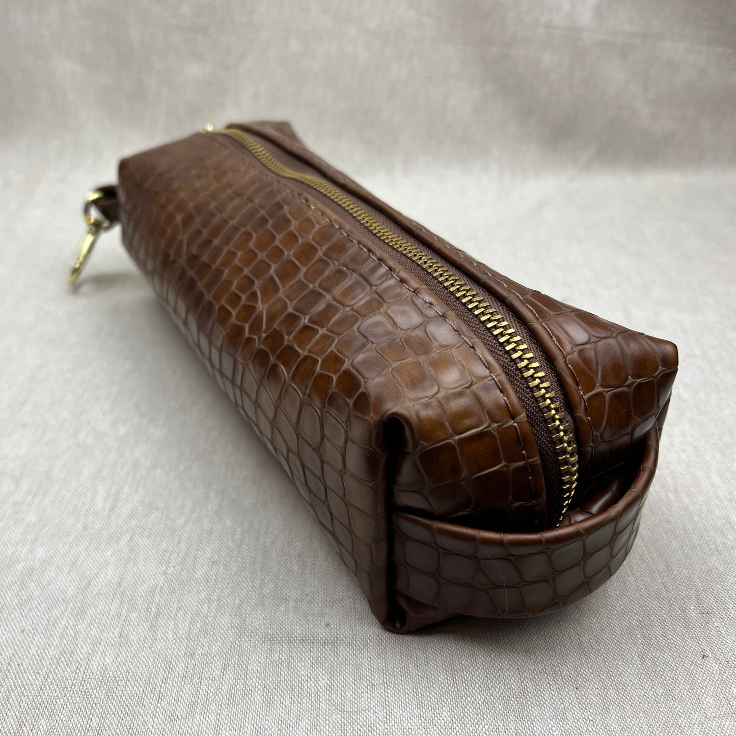 A brown crocodile-embossed leather zippered pouch with a gold-tone zipper and pull, shown on a light gray textured fabric background.