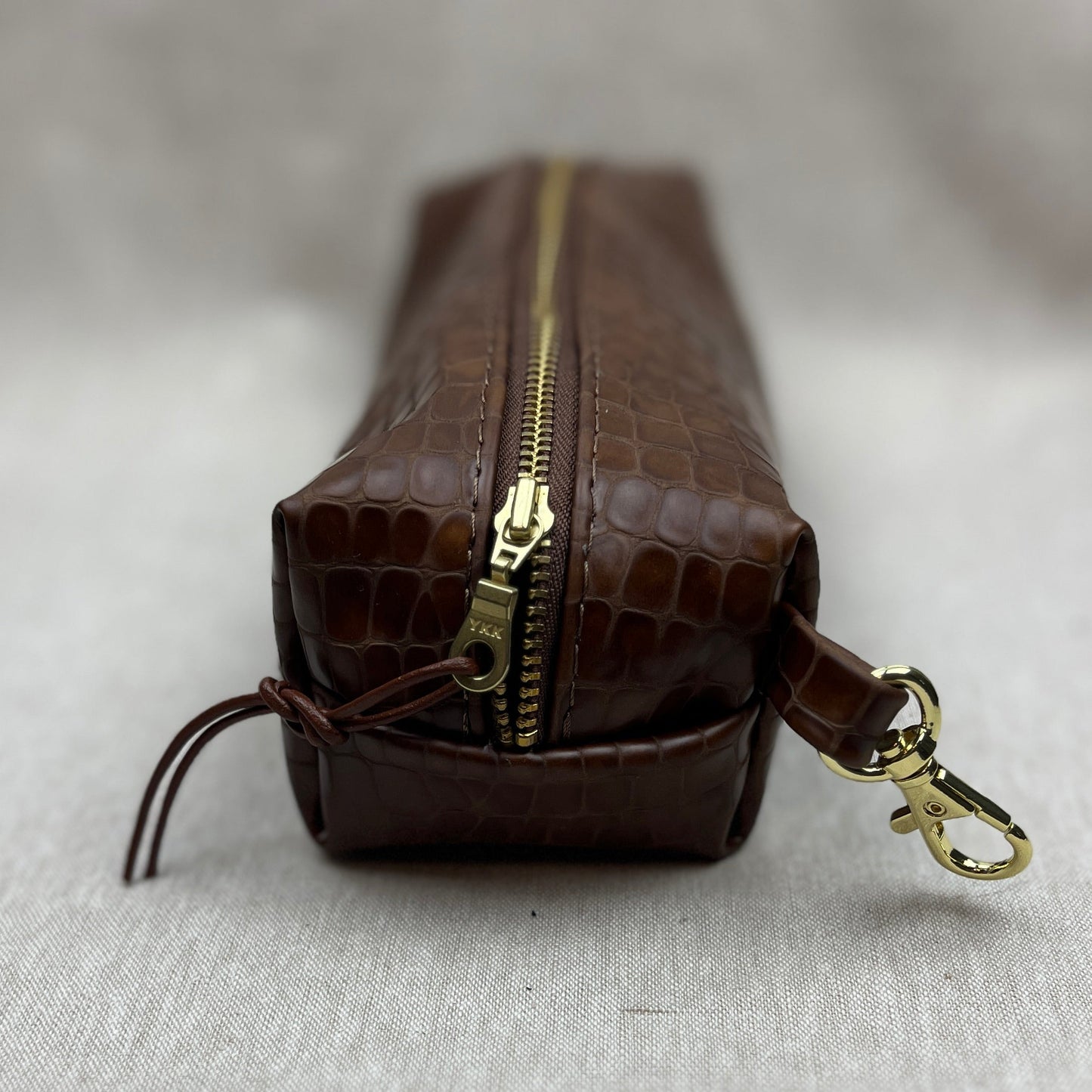 A brown crocodile-embossed leather zippered pouch with gold-tone hardware, shown from a low angle on a light gray textured fabric background.