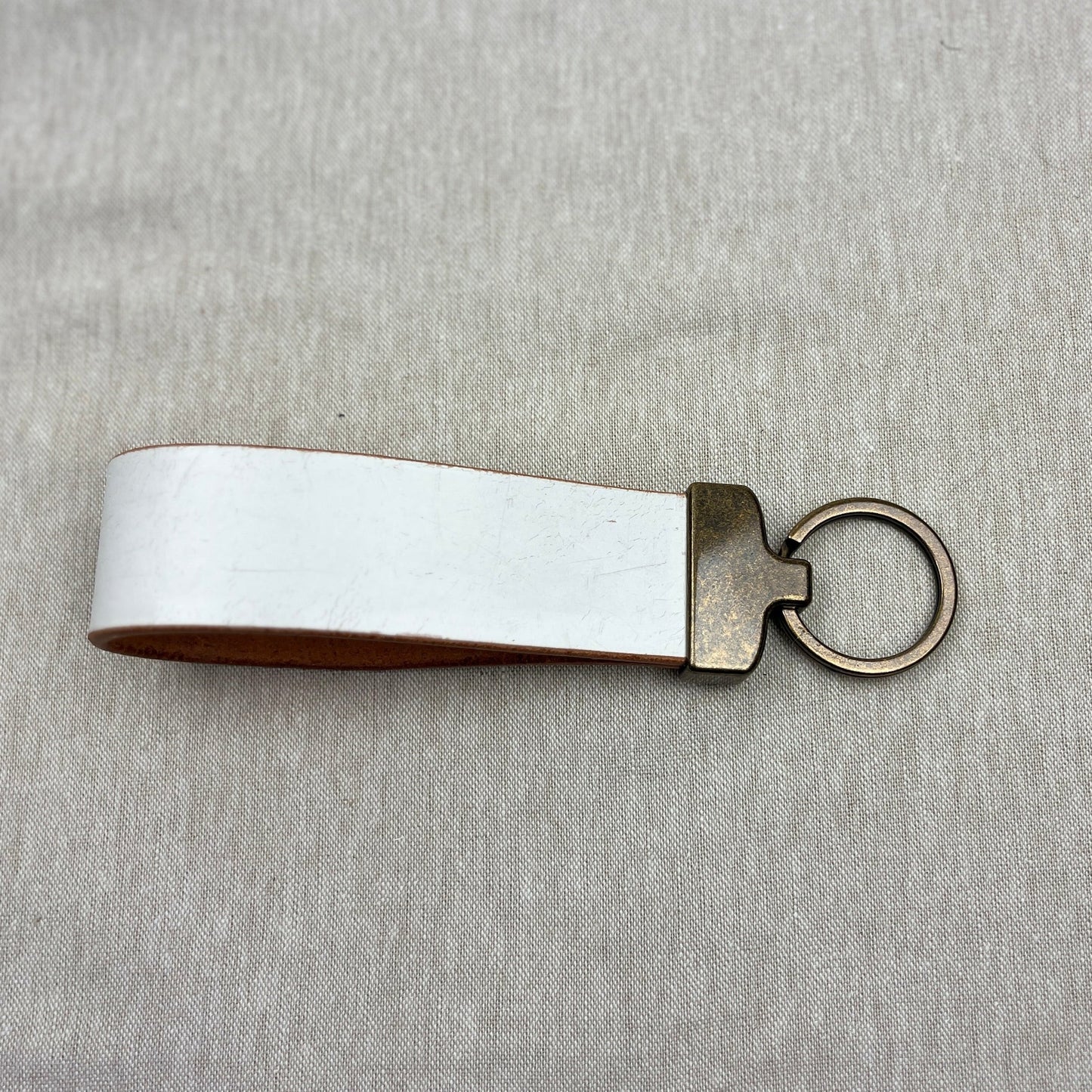 A blank white leather keychain loop with a bronze metal split ring and connector, displayed on a textured light gray fabric background.