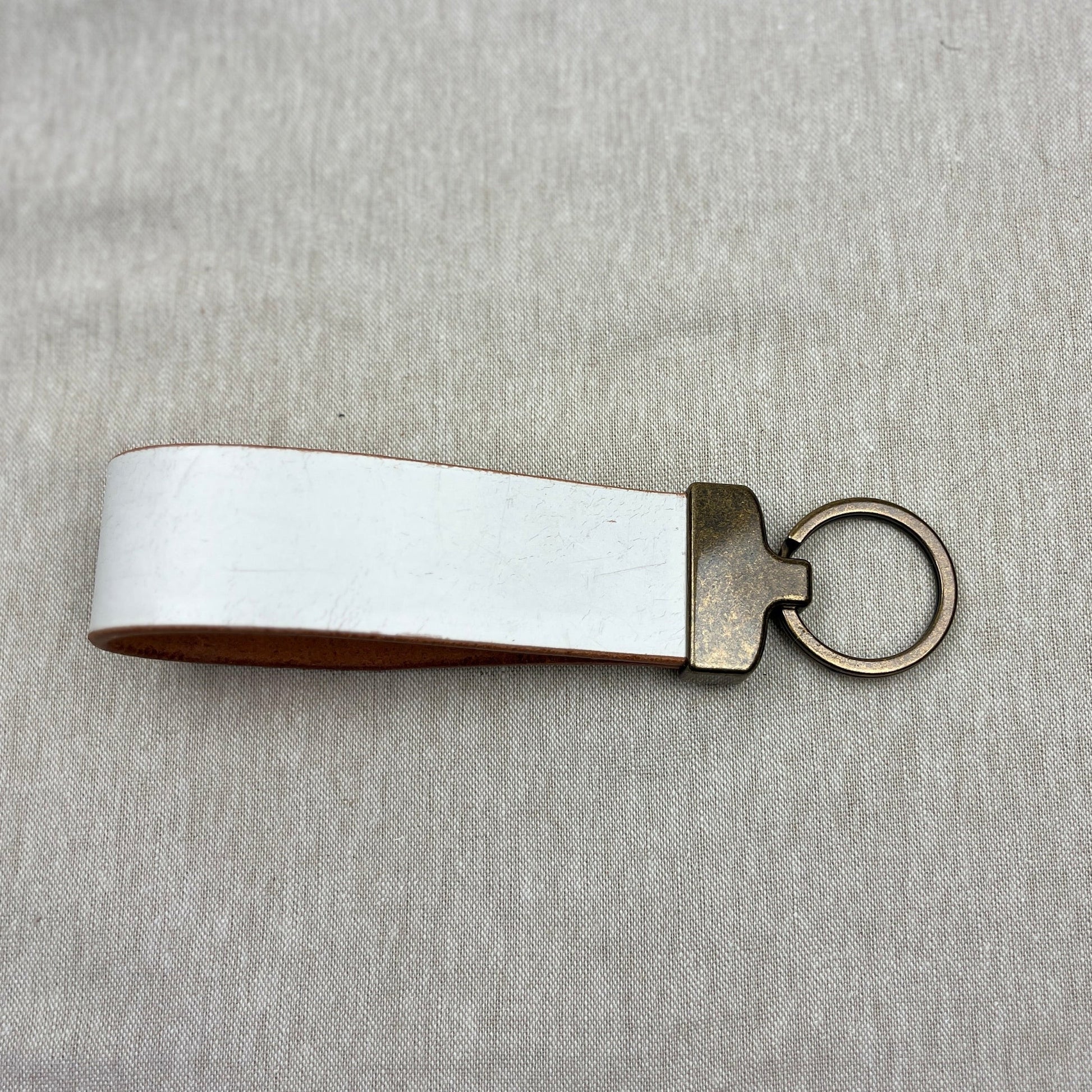 A blank white leather keychain loop with a bronze metal split ring and connector, displayed on a textured light gray fabric background.