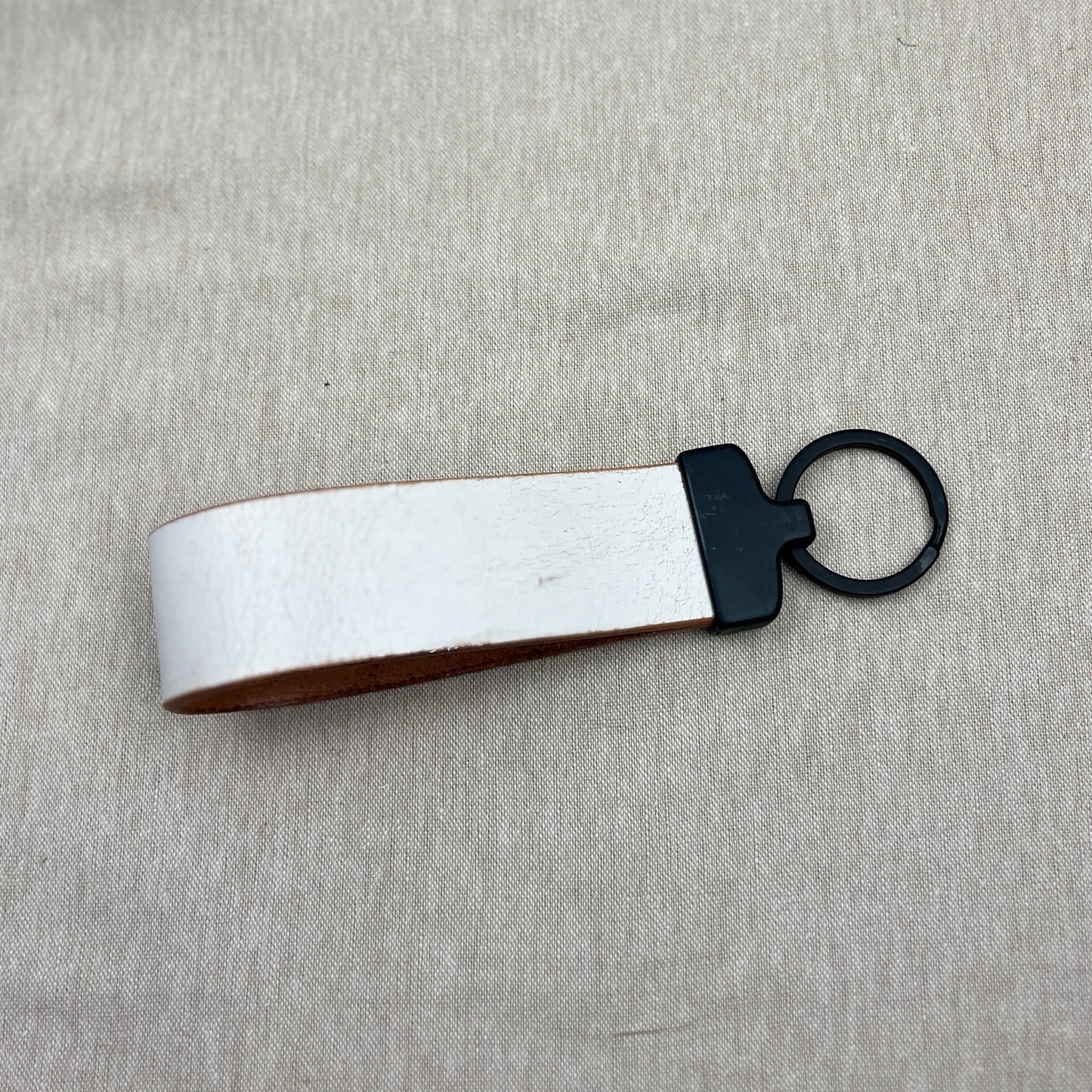 A leather keychain with a white exterior, brown interior, and black metal ring, placed on a light gray textured fabric background.