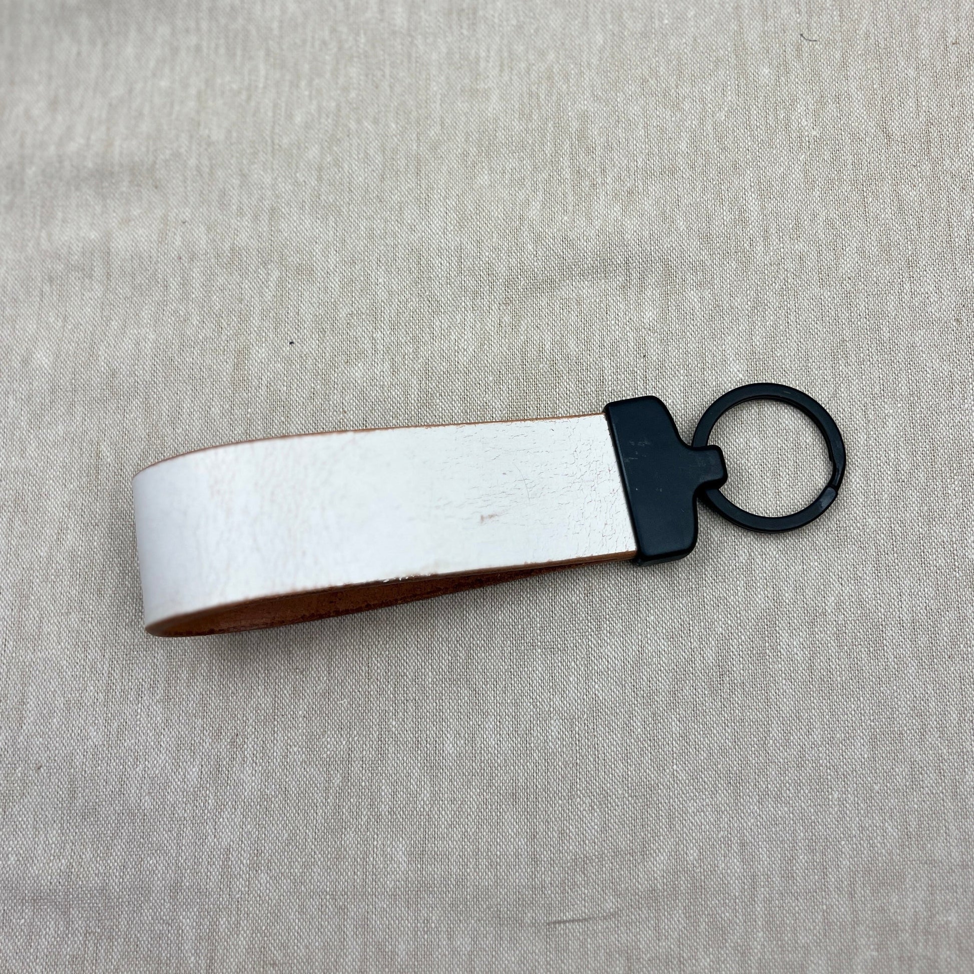 A leather keychain with a white exterior, brown interior, and black metal ring, placed on a light gray textured fabric background.