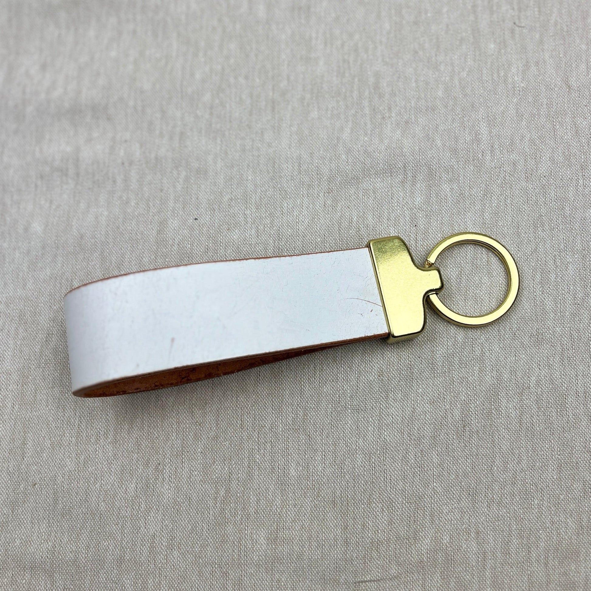 A white leather keychain strap with a gold-tone metal connector and key ring, shown on a textured light gray fabric background.