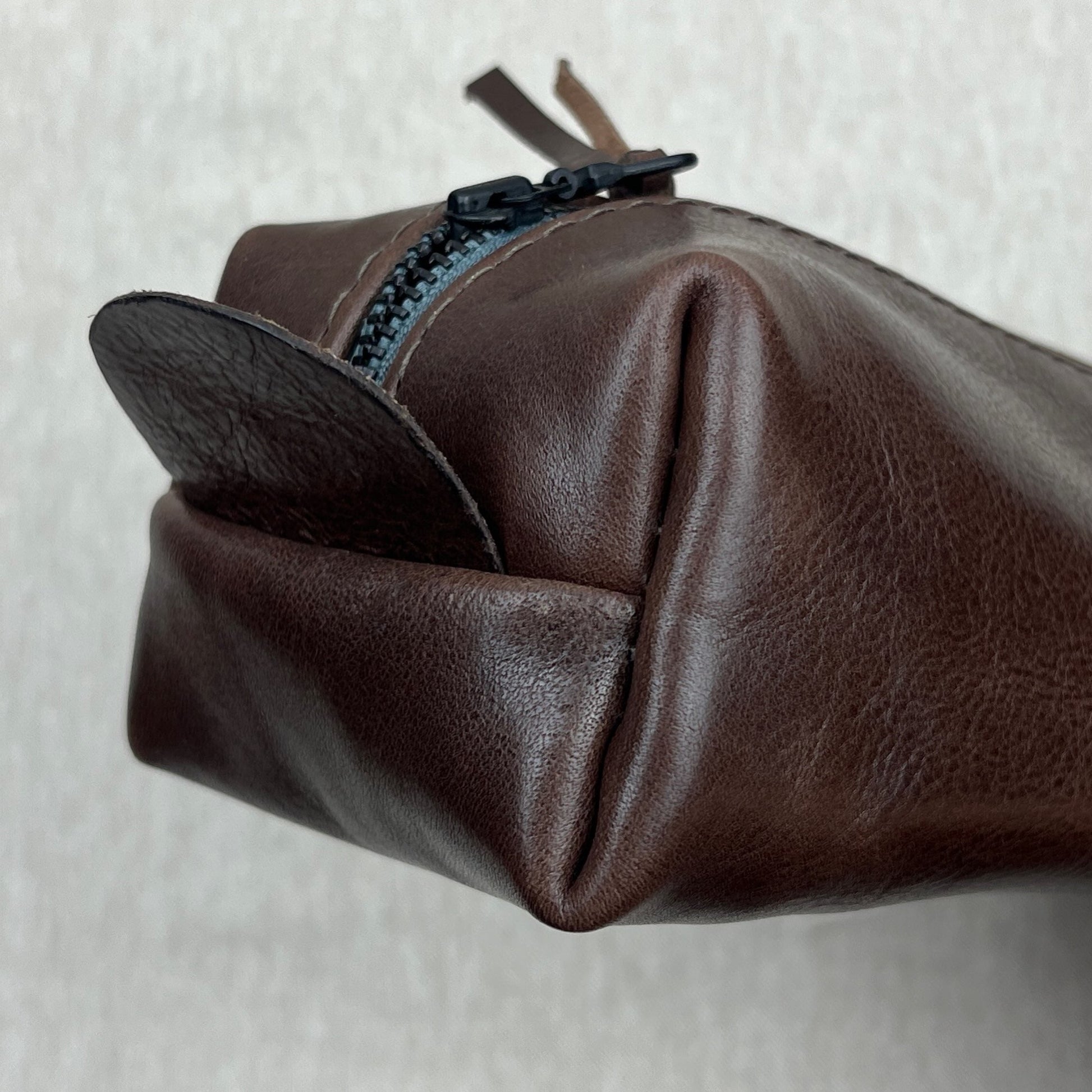 A close-up of a dark brown leather pouch or cosmetic bag, showing the textured leather material, a black zipper, and a matching leather pull tab against a light gray background.