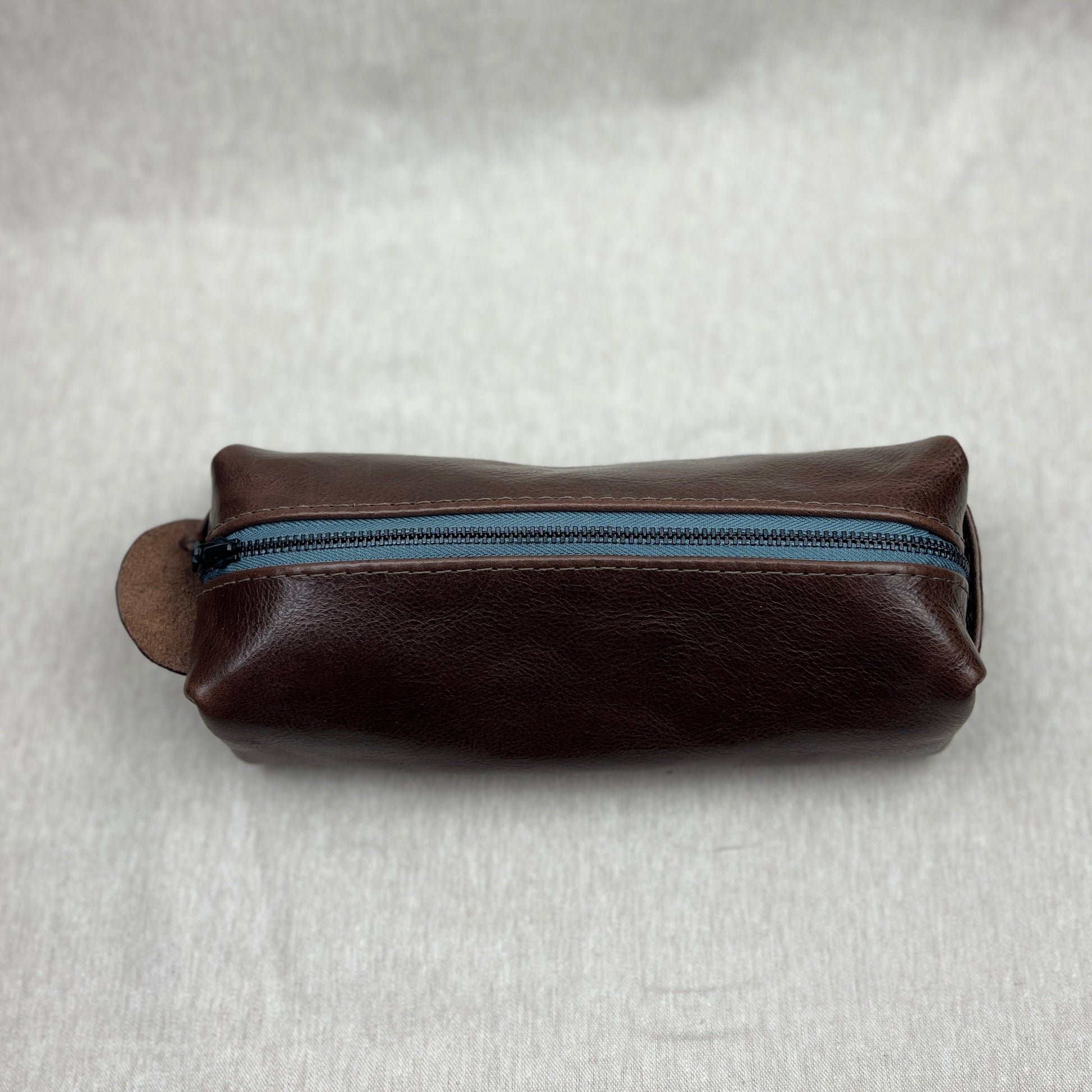 A dark brown leather zippered pouch with a blue zipper track, resting on a textured light gray fabric background.