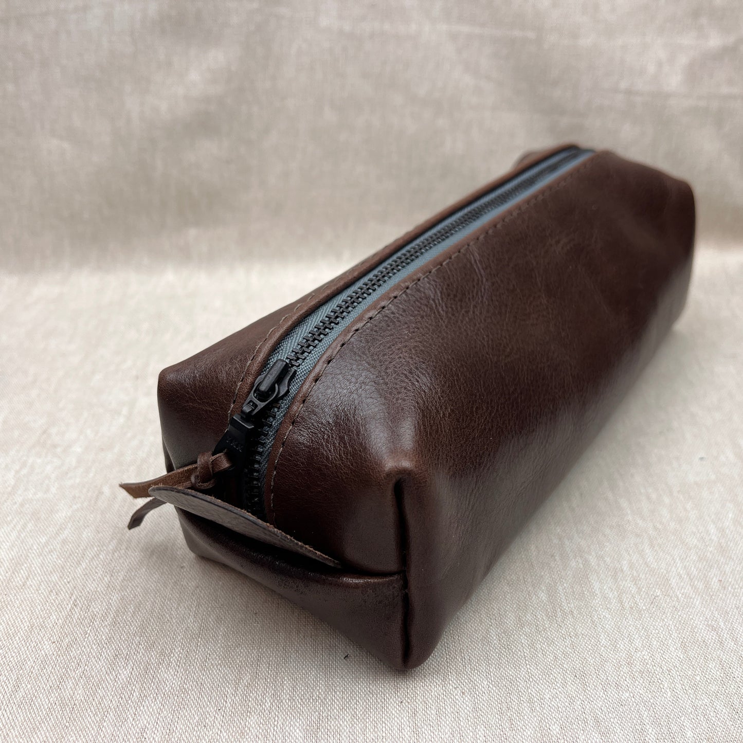A dark brown leather zippered pouch with a black zipper, shown on a textured light beige fabric background.