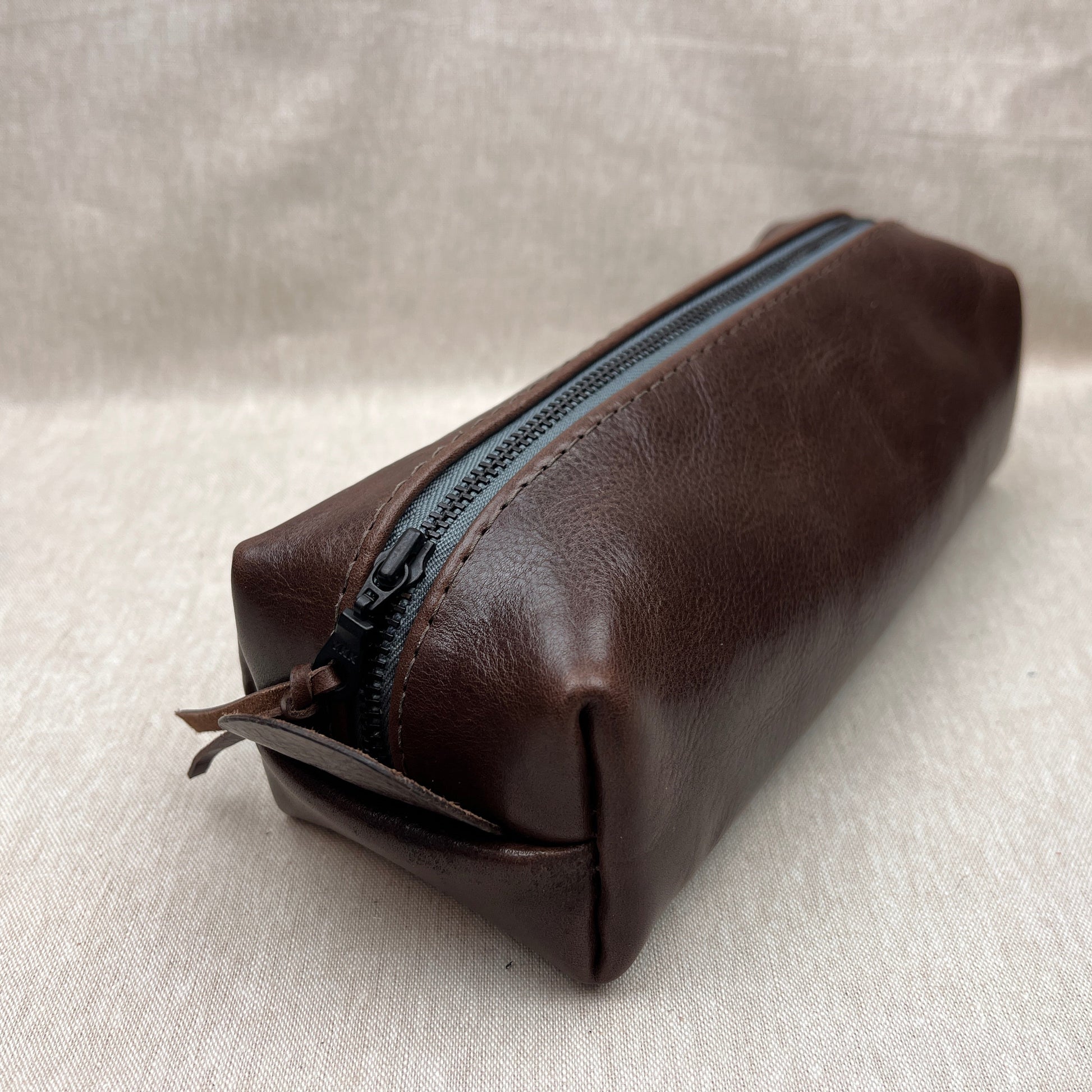 A dark brown leather zippered pouch with a black zipper, shown on a textured light beige fabric background.