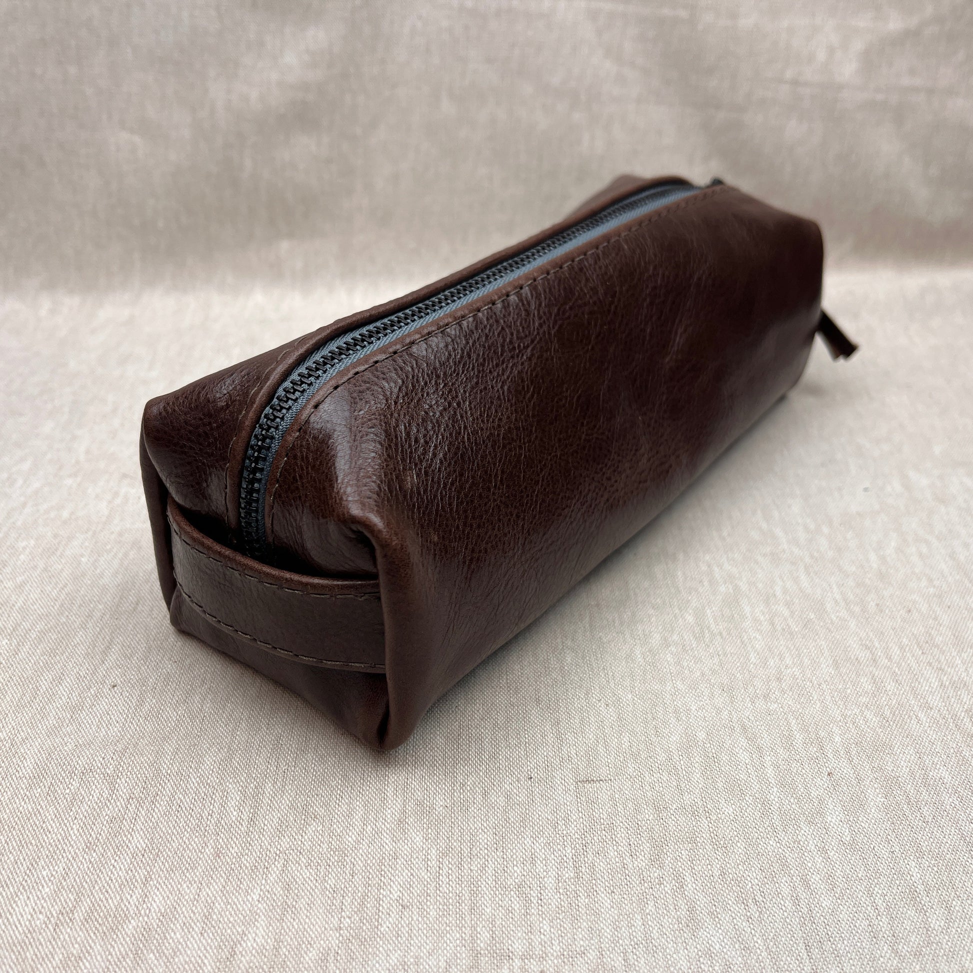 A dark brown leather zippered pouch with a blue zipper, resting on a light beige textured fabric background.
