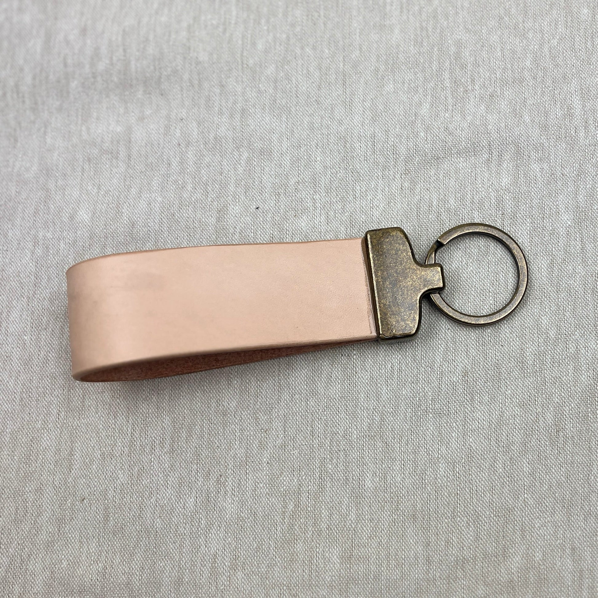 A light beige leather keychain strap with an antique bronze metal connector and key ring, shown on a textured gray background.