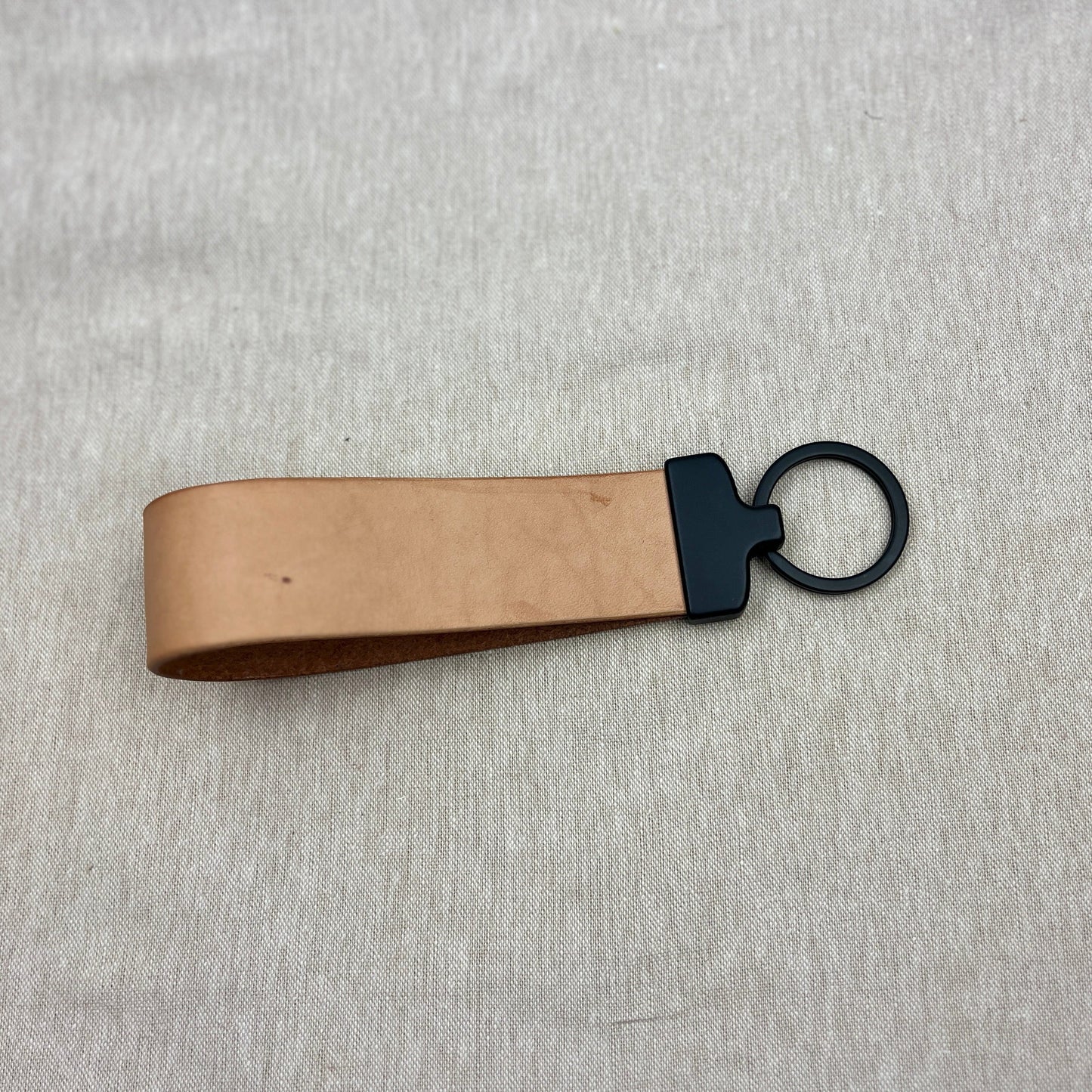 A tan leather keychain strap with a black metal connector and key ring, shown on a light gray textured fabric background.