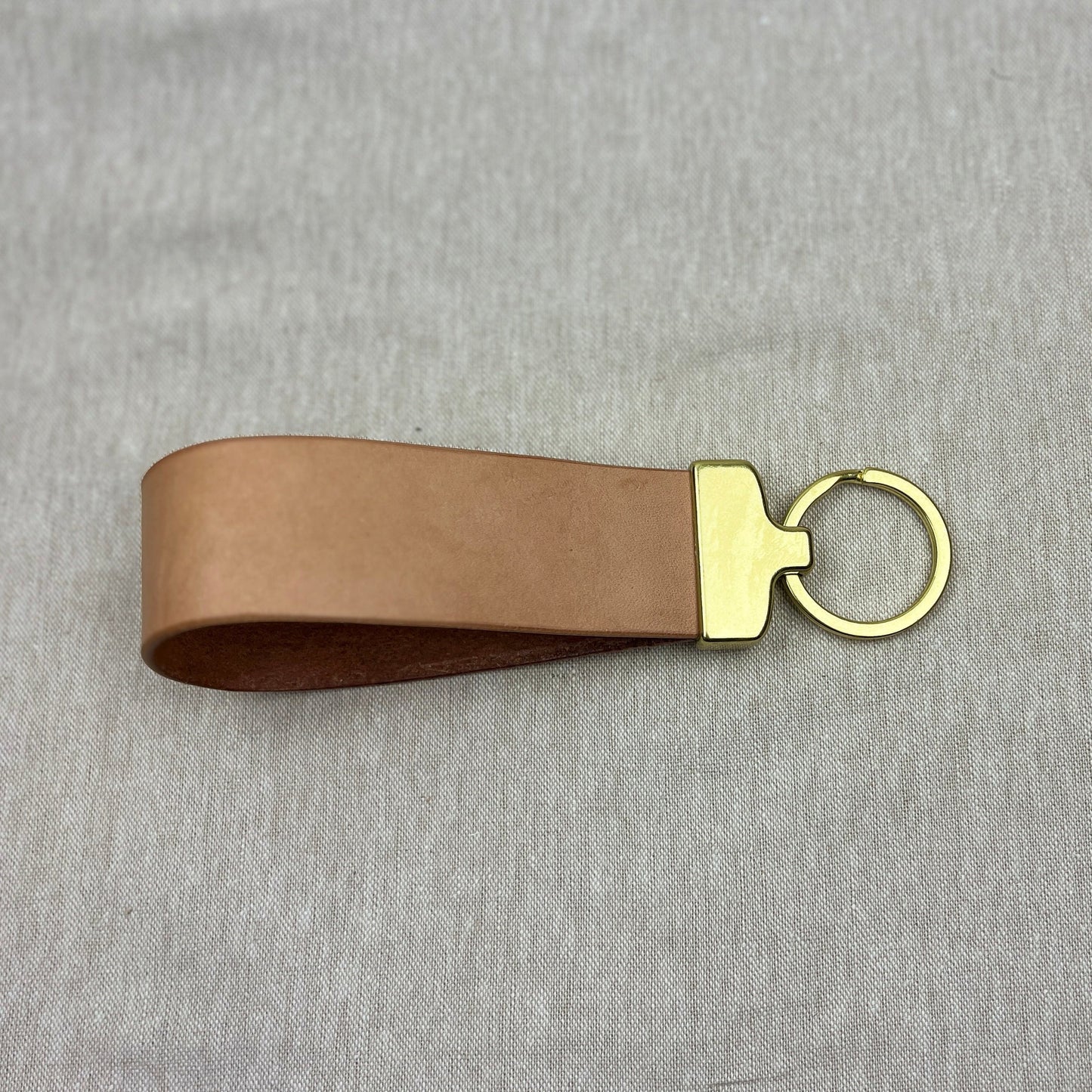 A light brown leather keychain strap with a gold-tone metal connector and key ring, displayed on a textured gray fabric background.