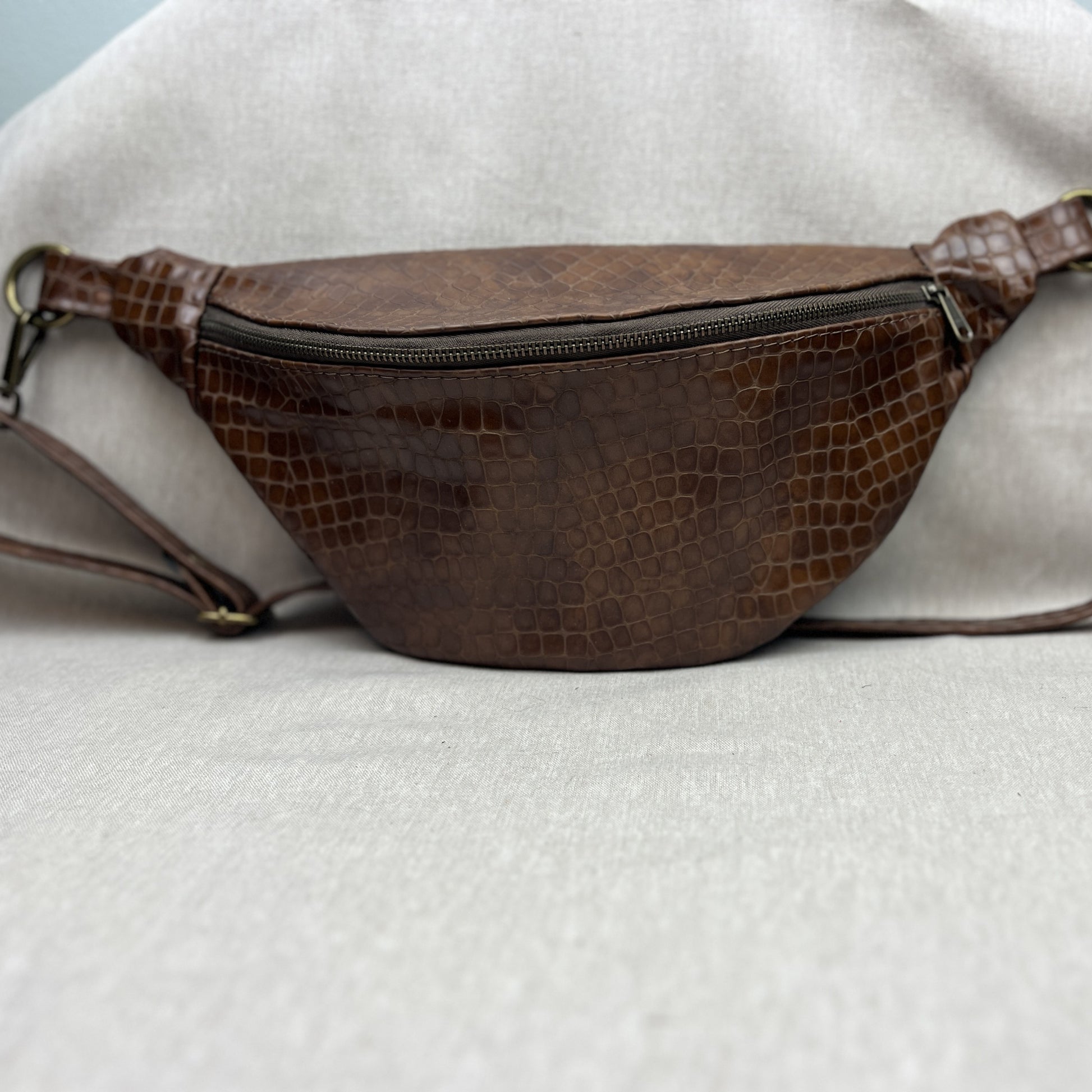 A brown faux-crocodile leather fanny pack with a zippered front pocket and adjustable strap, resting on a light gray fabric background.