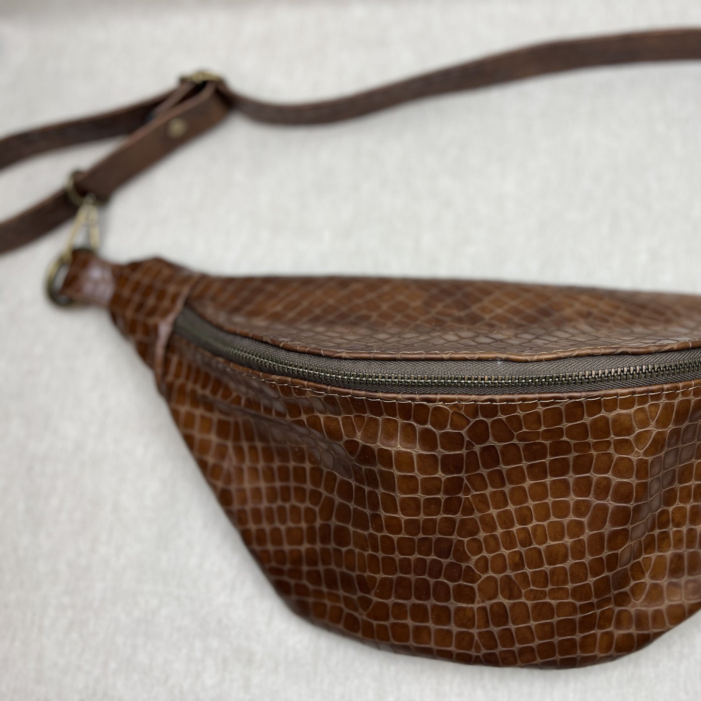 A brown faux-crocodile leather fanny pack with a matching adjustable shoulder strap, resting on a light gray textured fabric background.