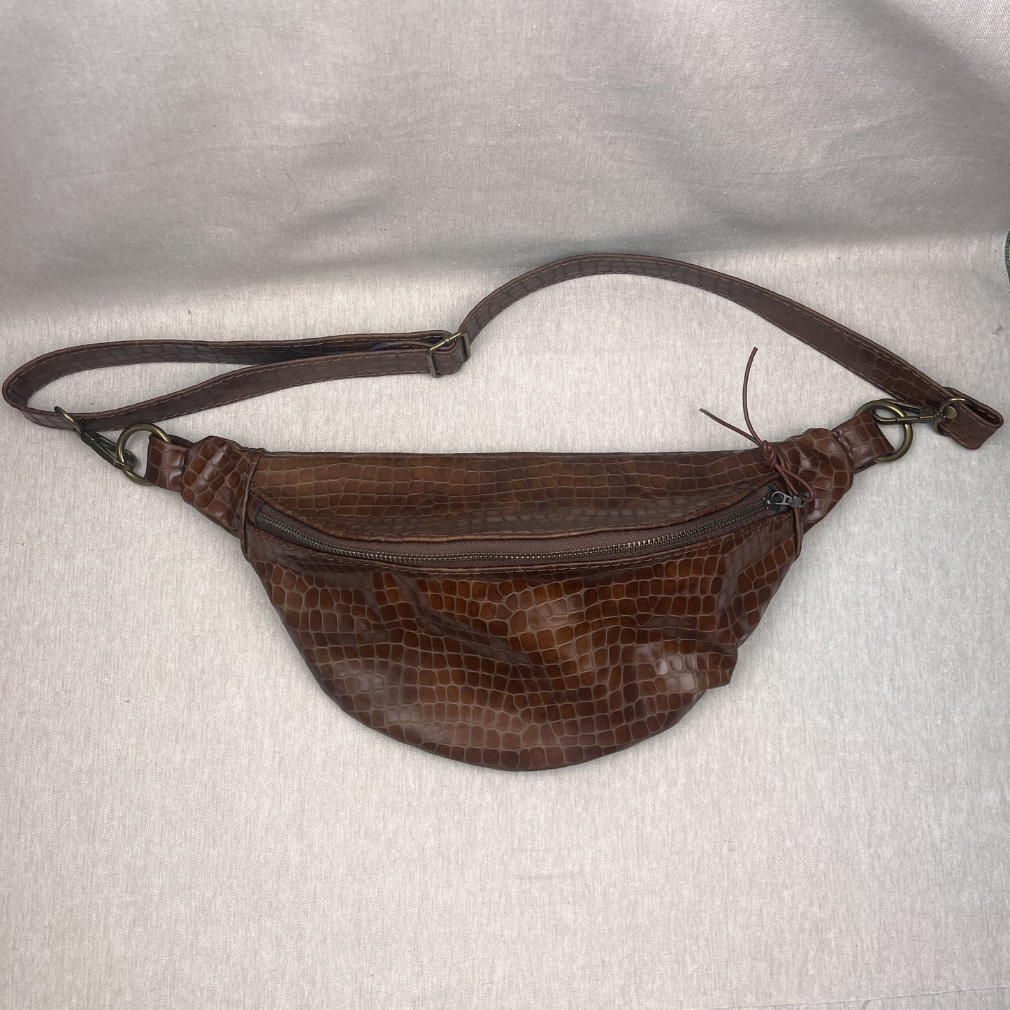 A brown crocodile-embossed leather fanny pack with a matching adjustable shoulder strap, laid flat on a light gray textured fabric background.