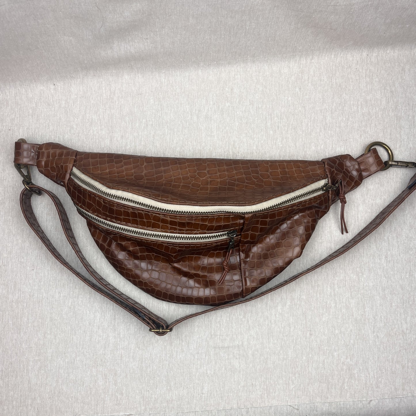 A brown faux-crocodile leather fanny pack with a textured pattern, featuring two silver zippers and a matching brown shoulder strap, laid flat on a light gray fabric background.