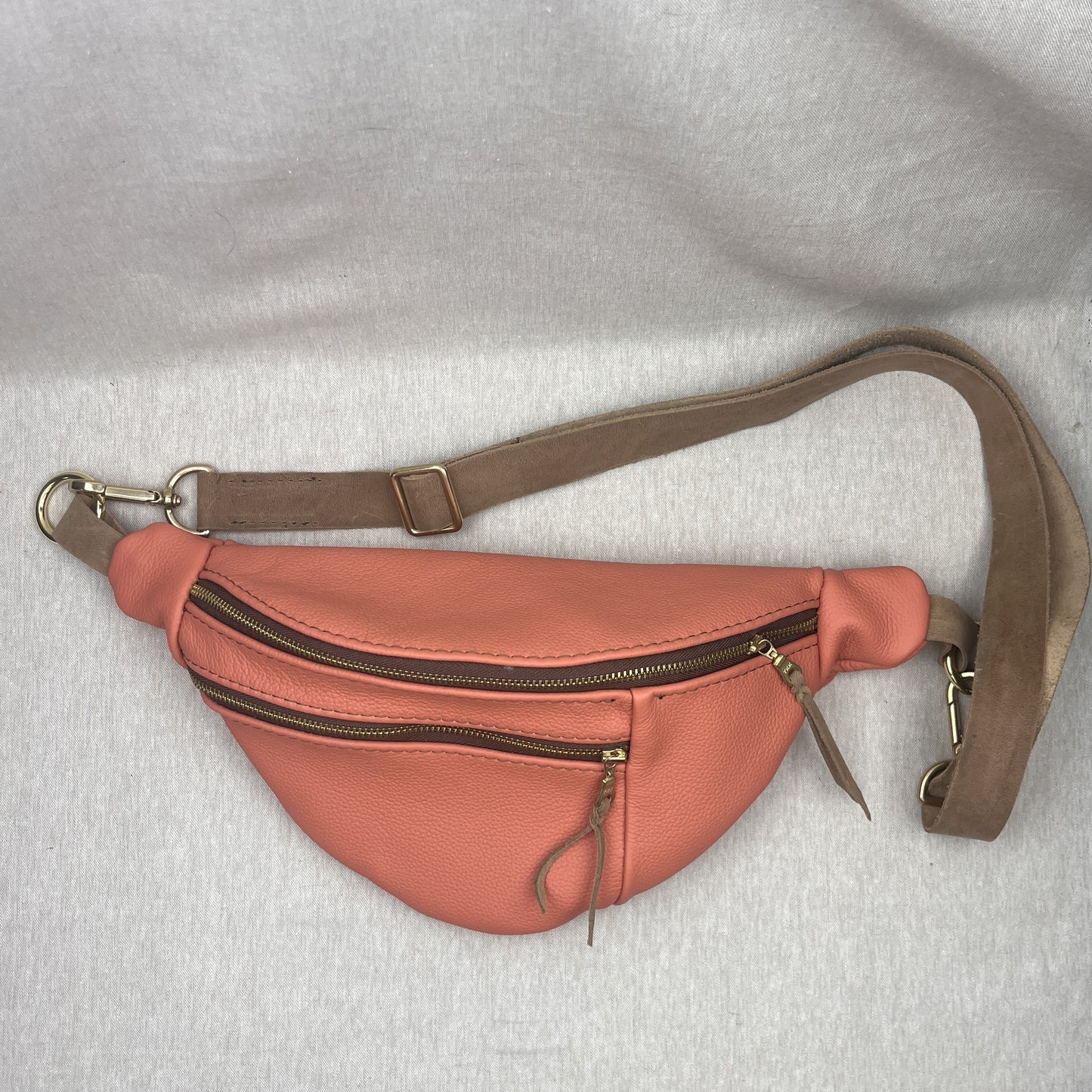 A coral pink faux-leather fanny pack with a brown adjustable strap and gold-tone hardware, laid flat on a light gray fabric background.