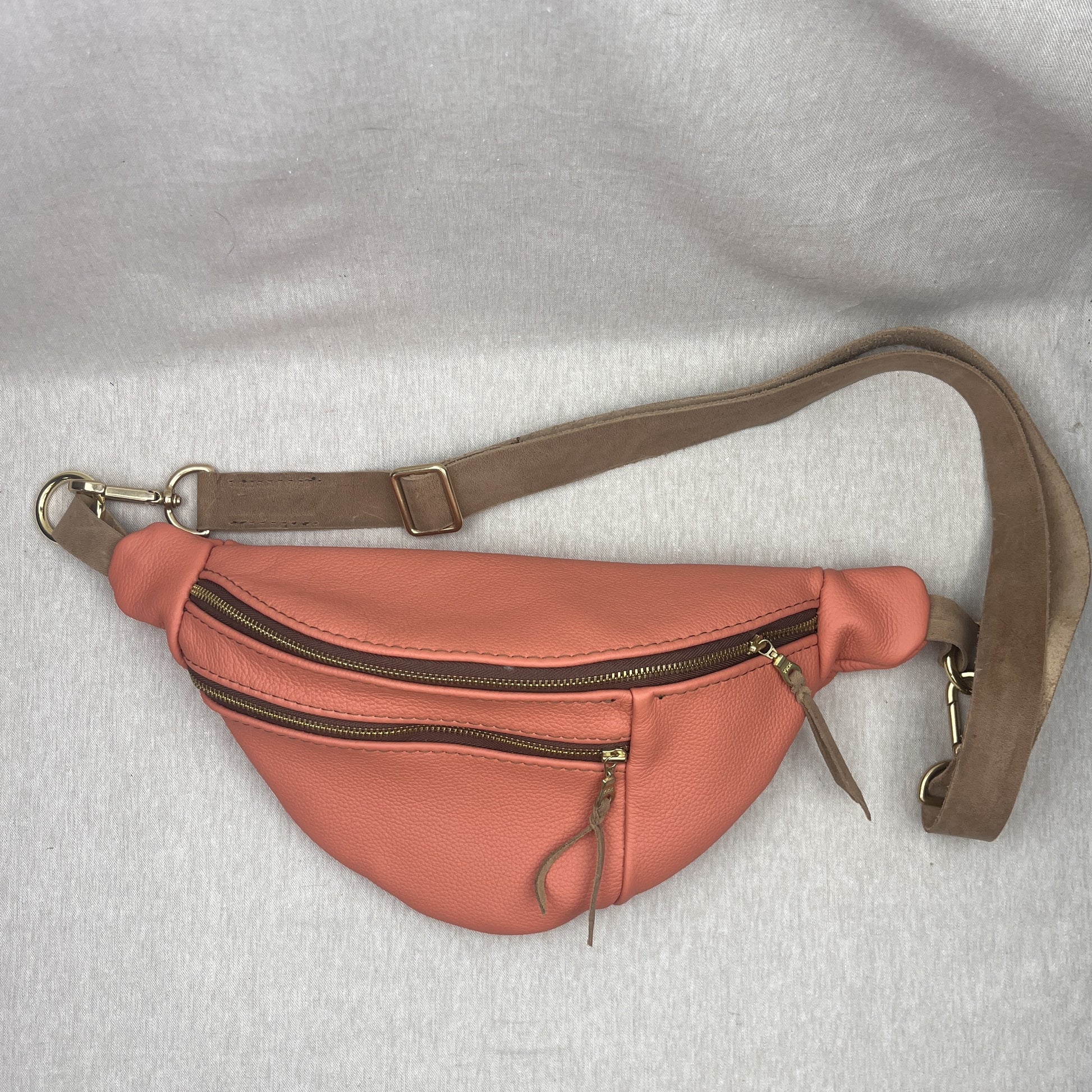 A coral pink faux-leather fanny pack with a brown adjustable strap and gold-tone hardware, laid flat on a light gray fabric background.