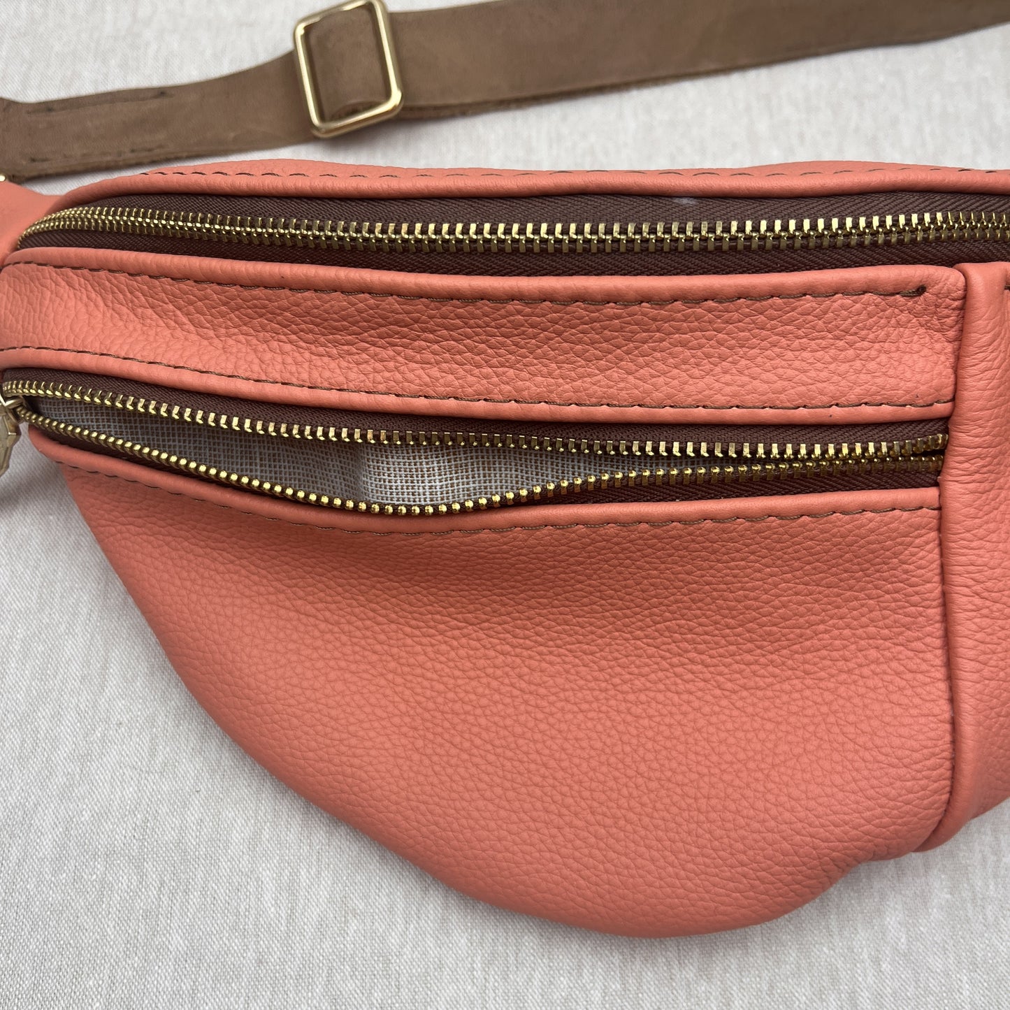 A close-up of a peach-colored pebbled leather fanny pack with gold-tone zippers and a brown strap on a light gray fabric background.