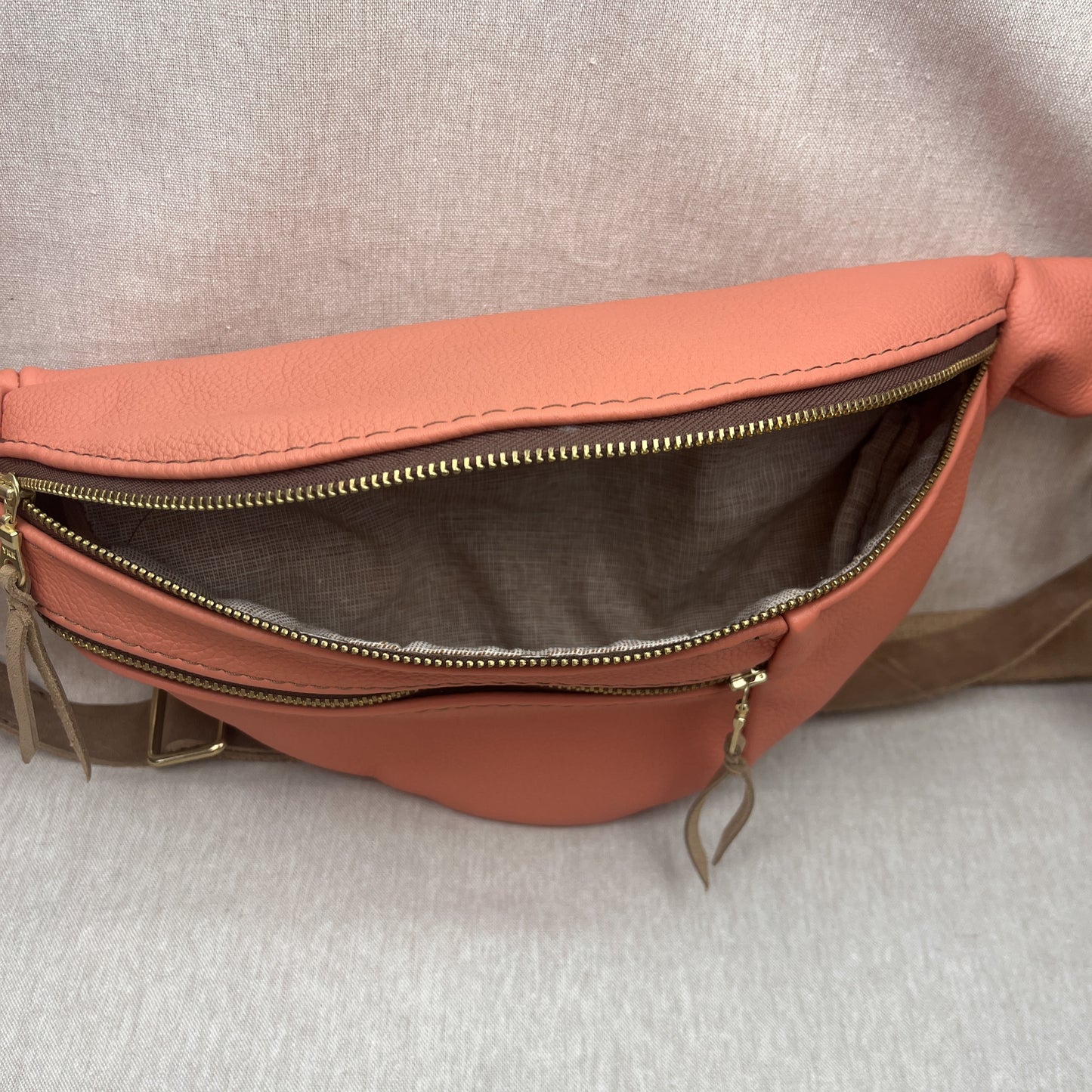 A peach-colored leather fanny pack shown open to display its interior, featuring a gold-tone zipper and a brown fabric lining, set against a neutral gray textured background.