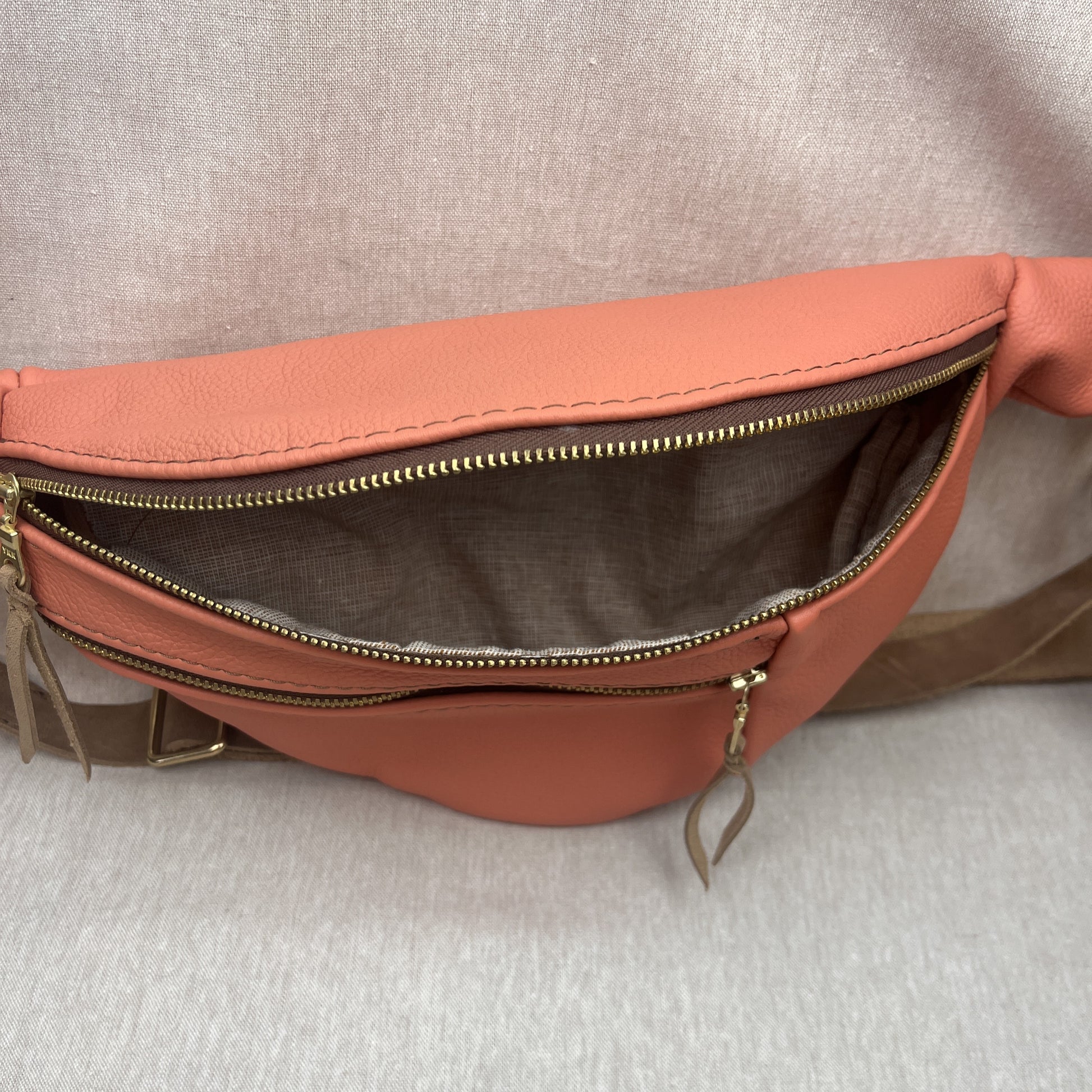 A peach-colored leather fanny pack shown open to display its interior, featuring a gold-tone zipper and a brown fabric lining, set against a neutral gray textured background.