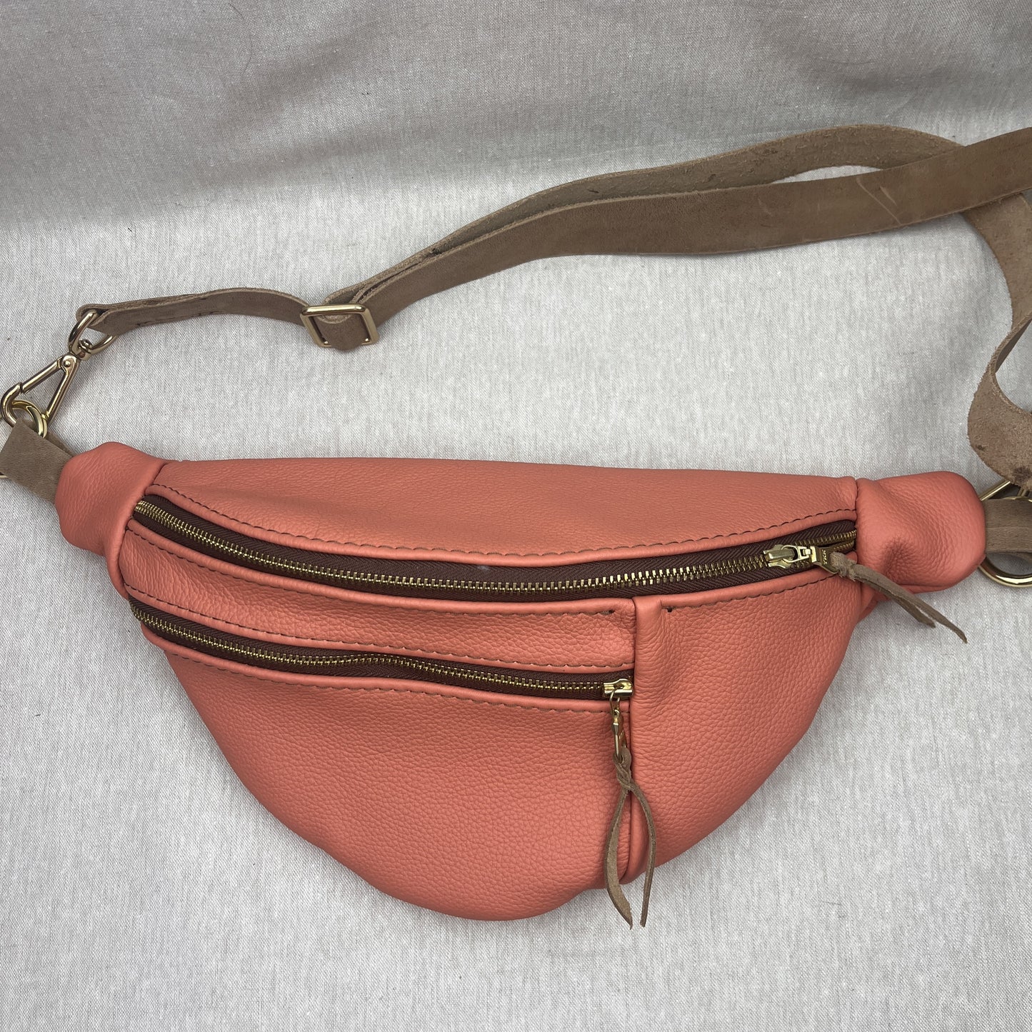 A peach-colored pebbled leather fanny pack with a brown adjustable strap and gold-tone hardware, laid flat on a light gray fabric background.