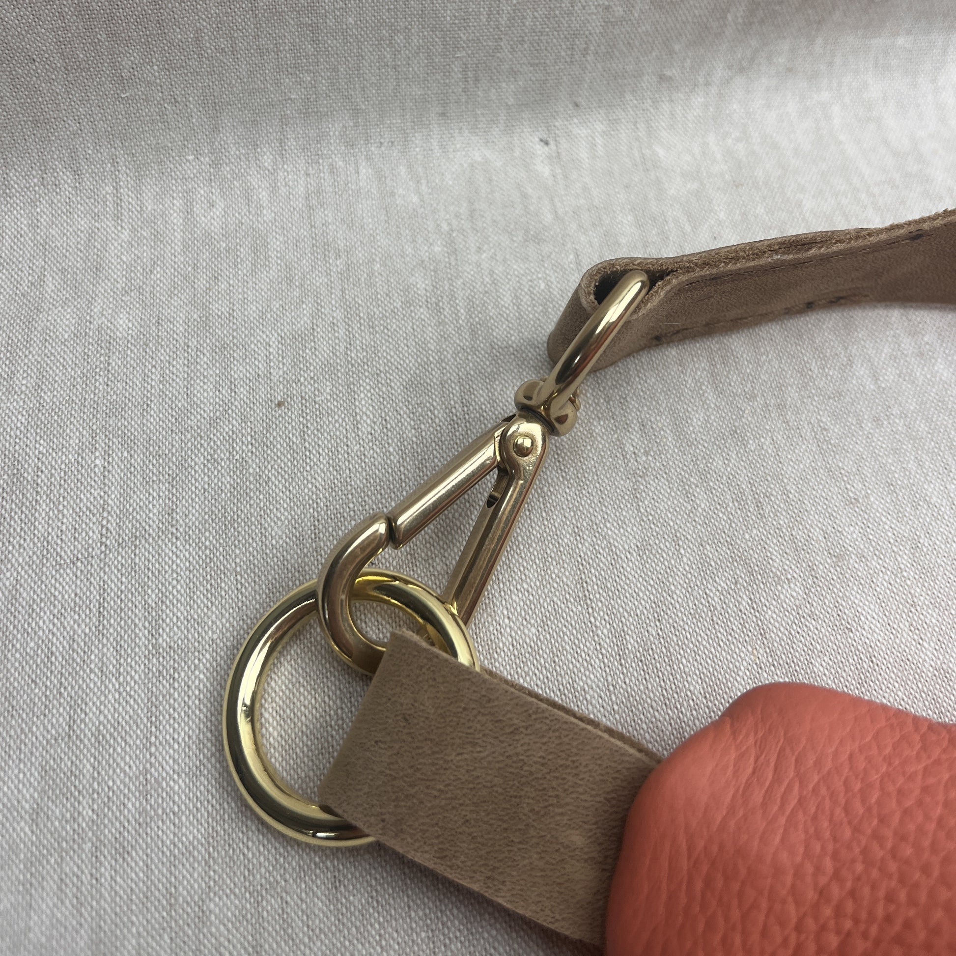 Close-up of a taupe leather strap attached to a gold-tone metal swivel snap hook and D-ring, with a finger holding the strap at the bottom right against a light gray textured fabric background.