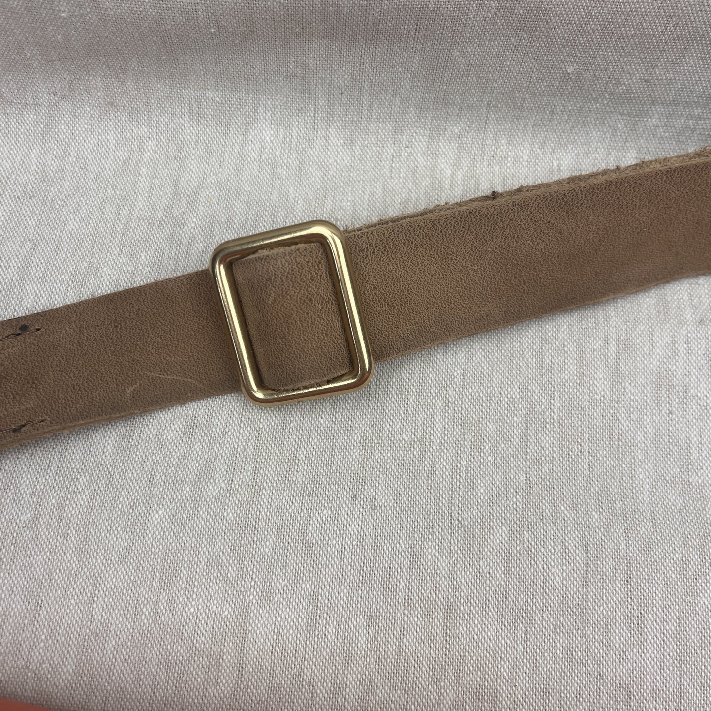 A close-up of a brown leather strap with a square gold-toned metal buckle, laid diagonally on a light gray textured fabric background.